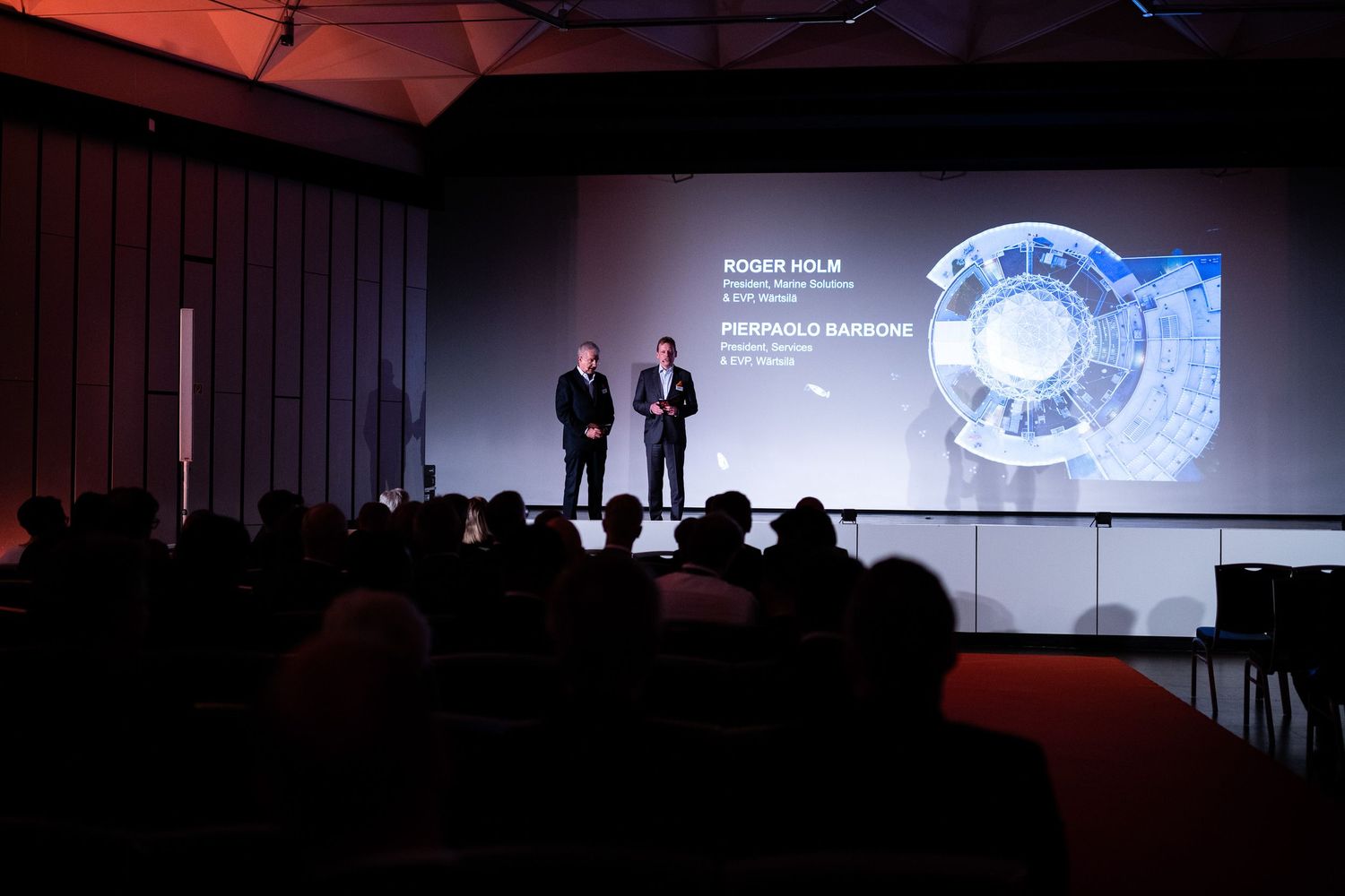 A dark conference room with presentation screens displaying technical information and data visualizations.