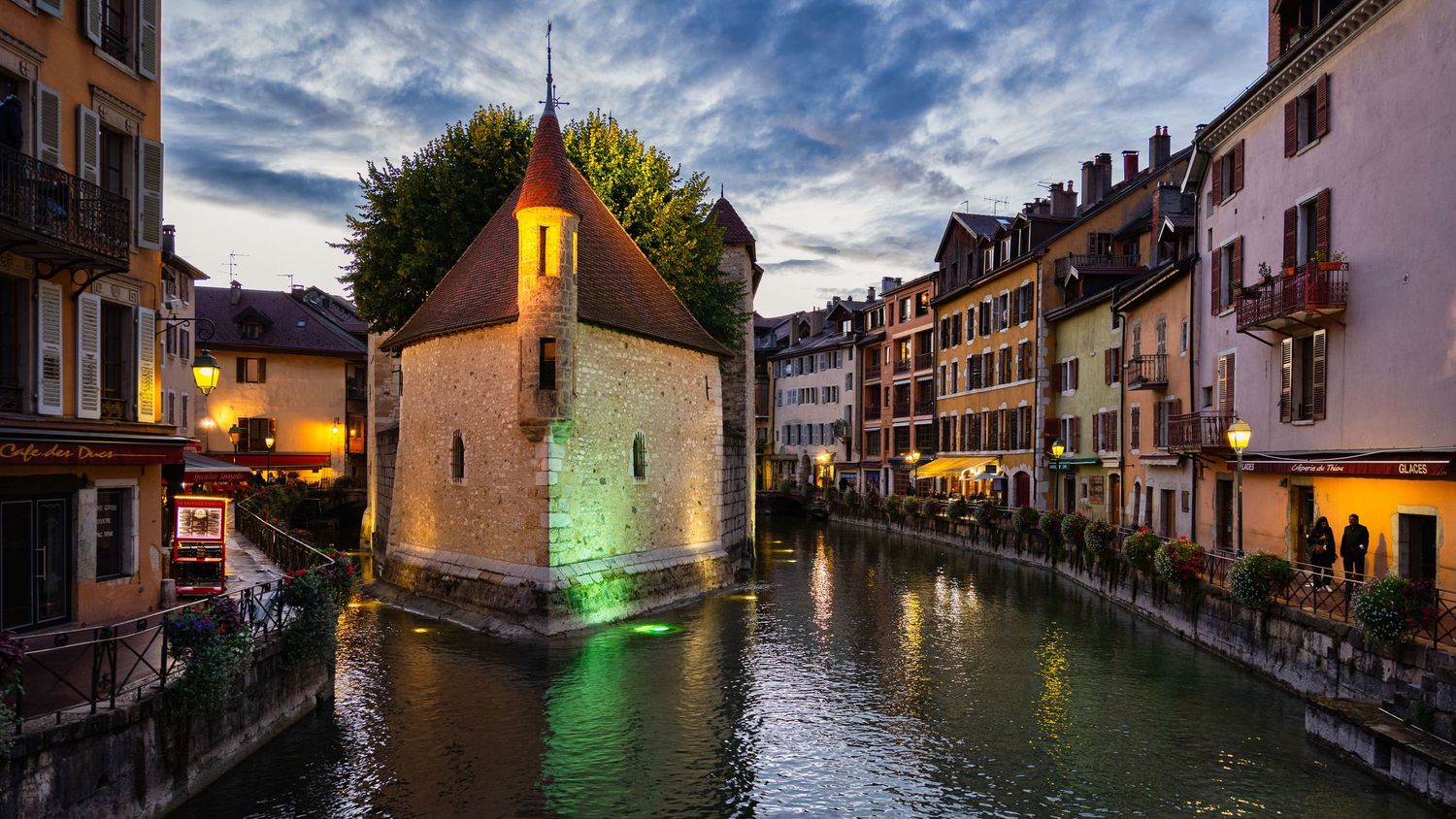 Night panorama of historic buildings reflecting in Annecy's canal with colorful lights.
