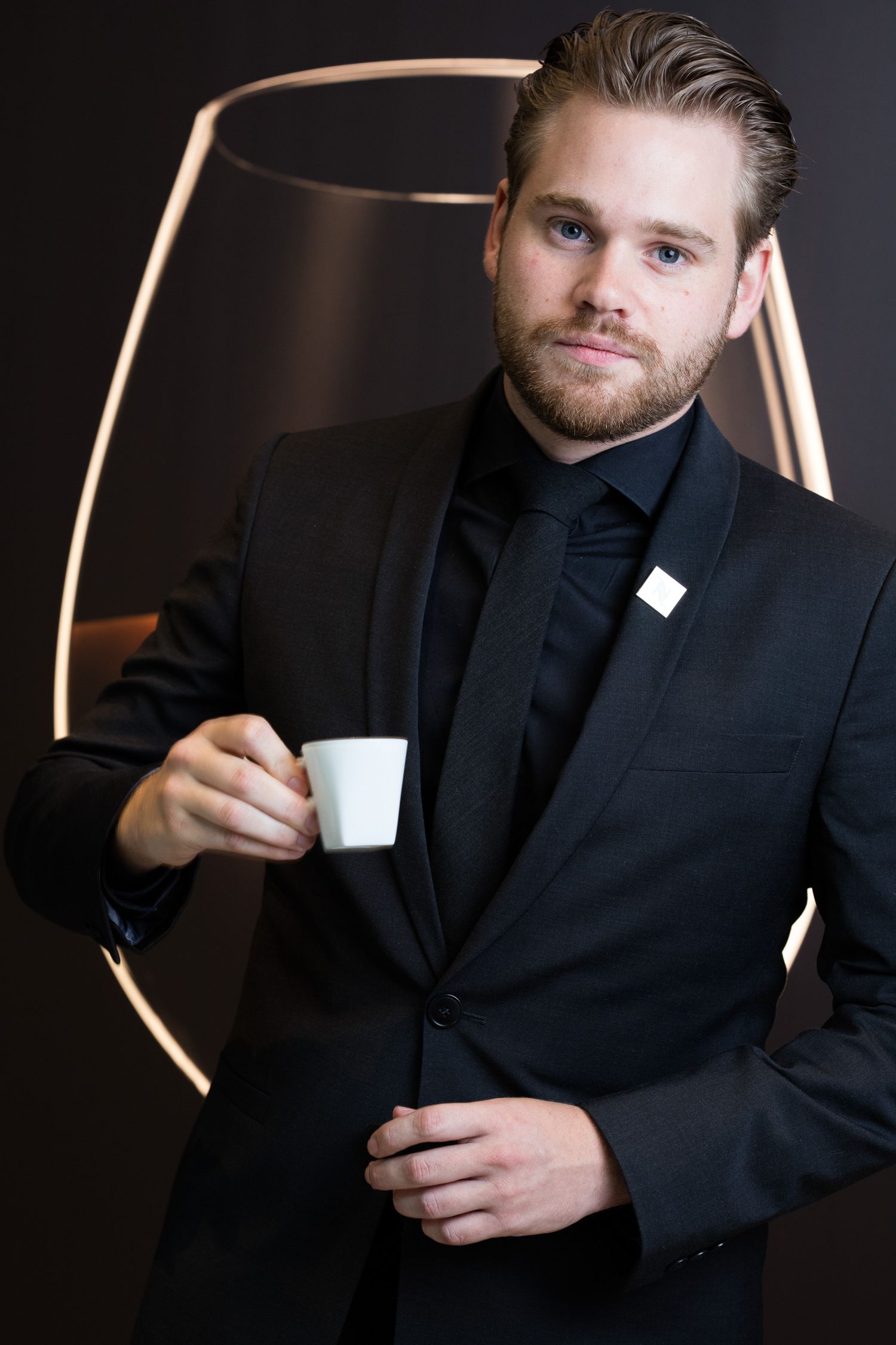 Professional portrait of someone in a black suit holding a white coffee cup against a dark background.