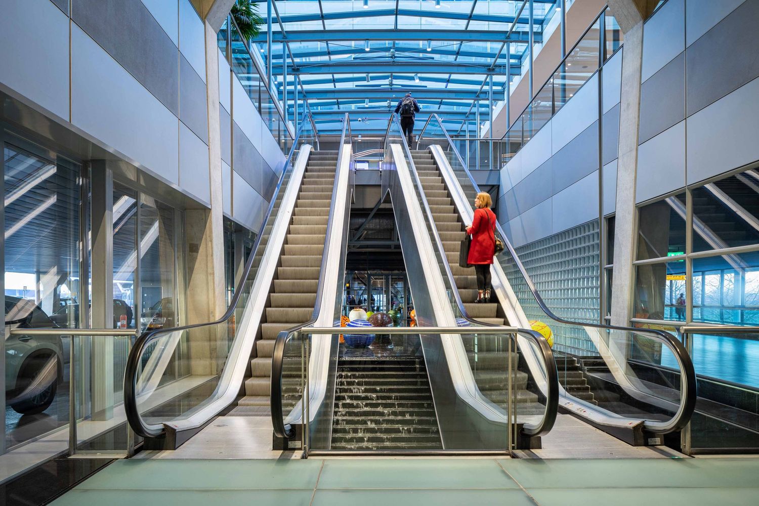 Modern escalators and stairs in a bright blue-tinted shopping mall with glass ceiling panels.