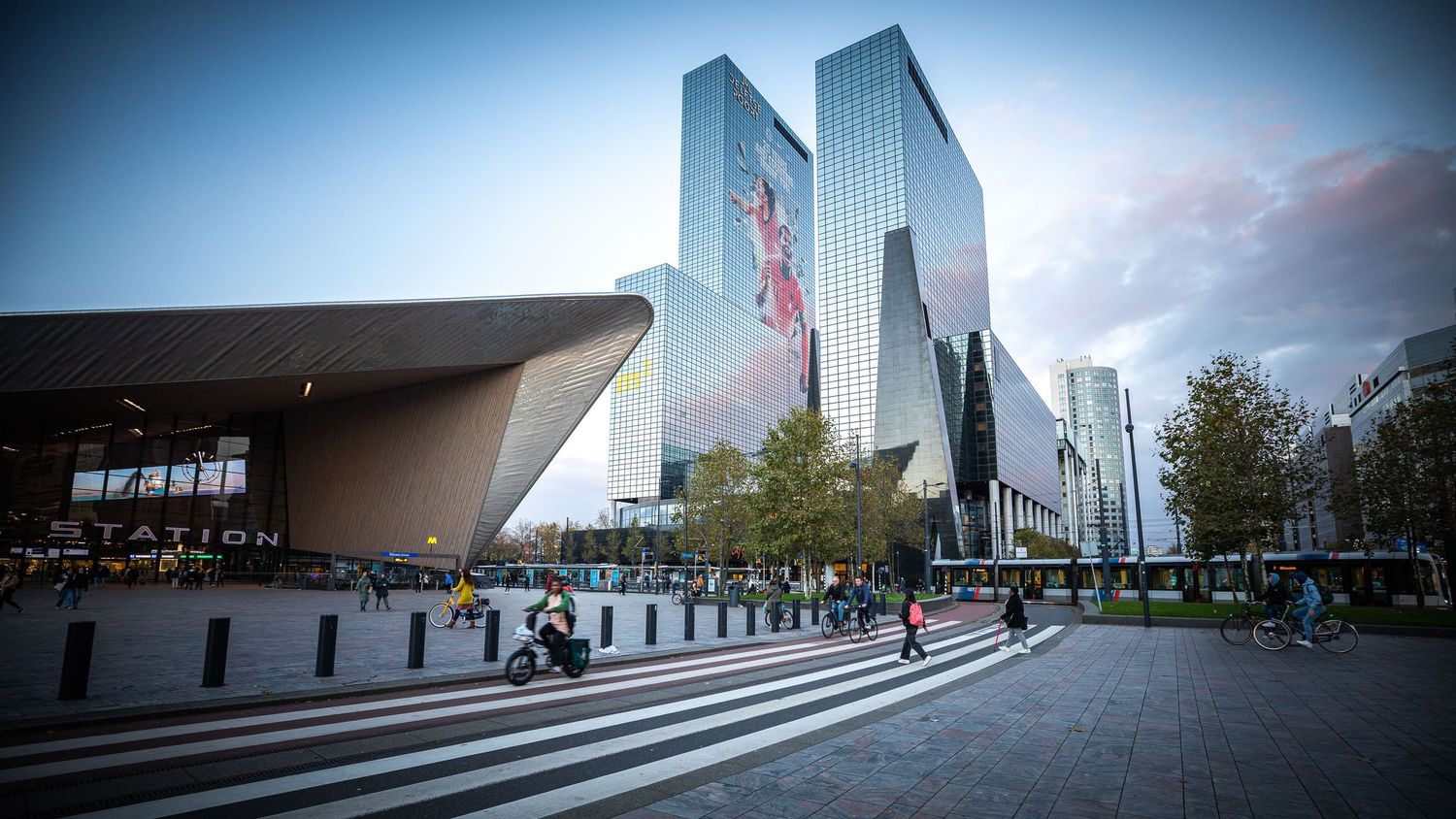 Modern Rotterdam Centraal Station with glass skyscrapers, pedestrians crossing zebra crossing at dusk.