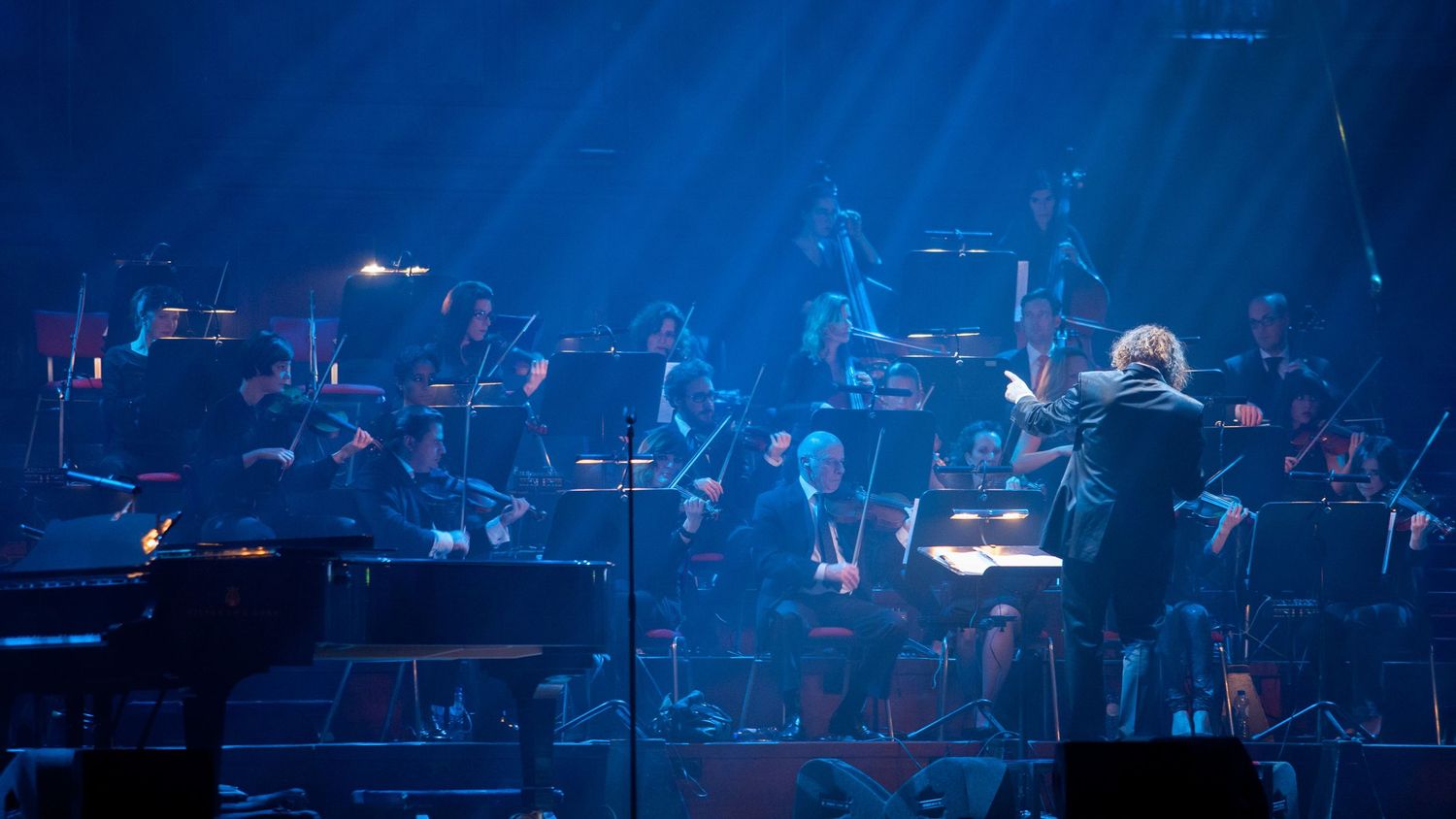 Orchestra performing on stage under dramatic blue lighting with conductor leading musicians in a concert hall.