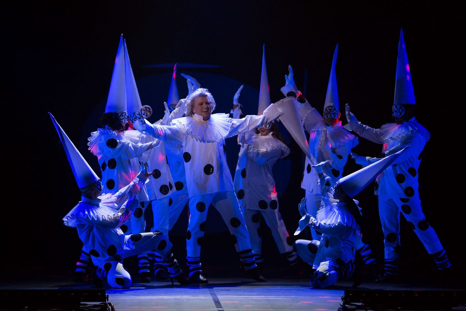 Performers in white polka-dot clown costumes and pointed hats dance on a dark stage under blue dramatic lighting.