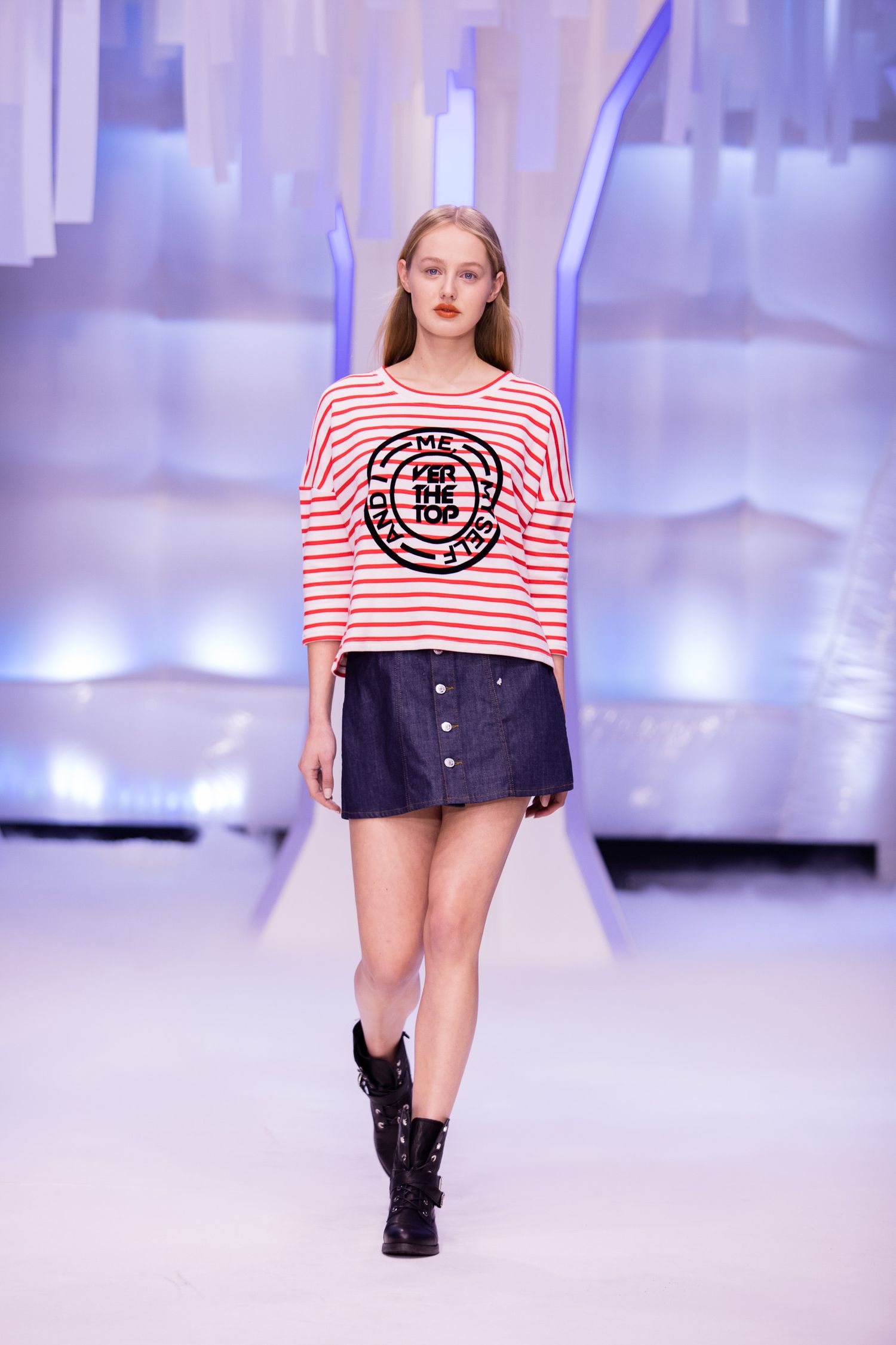 Model walks runway in red striped top with graphic print and navy mini skirt paired with black ankle boots.
