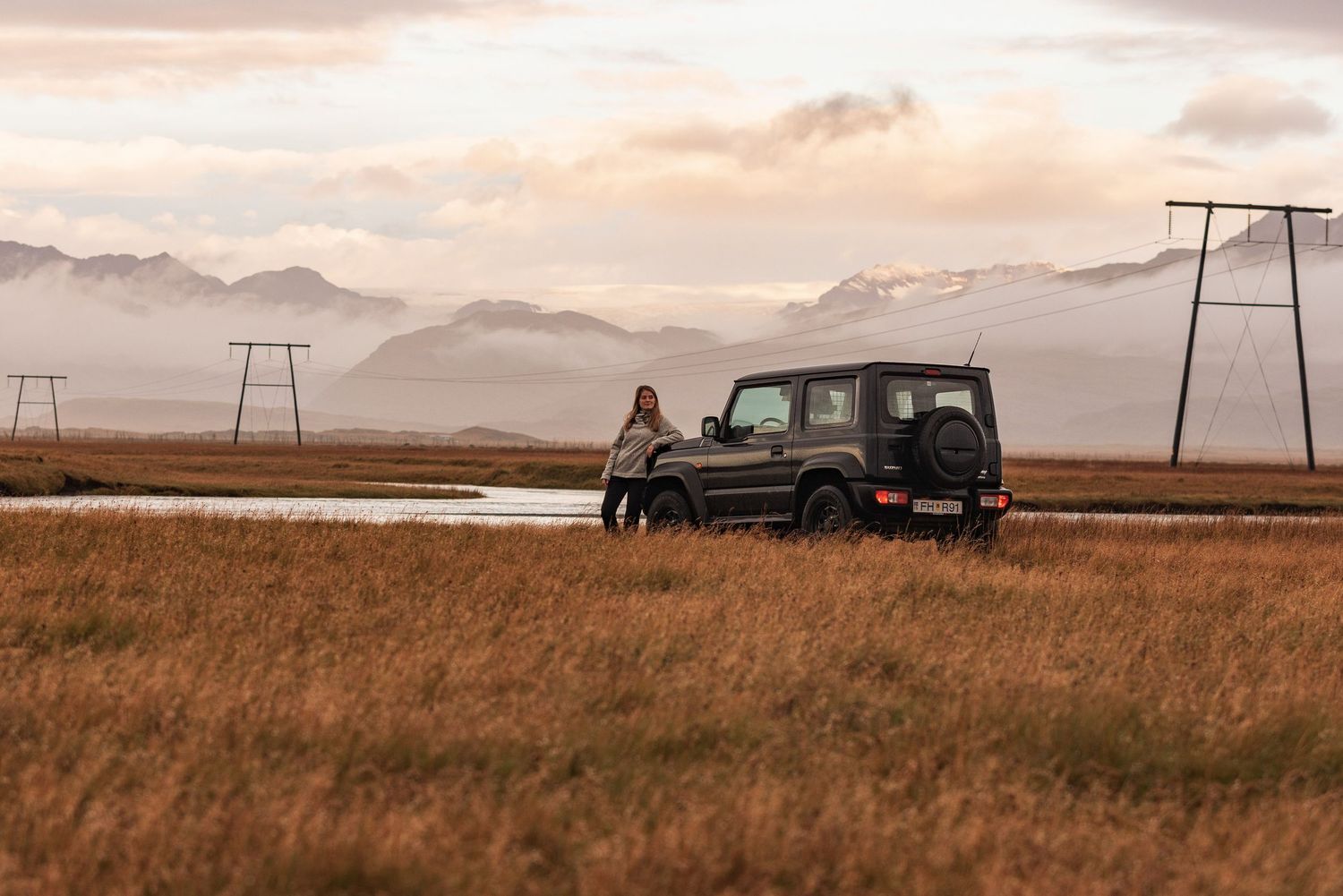 Black SUV parked in golden grassland with power lines and misty mountains in the background at sunset.
