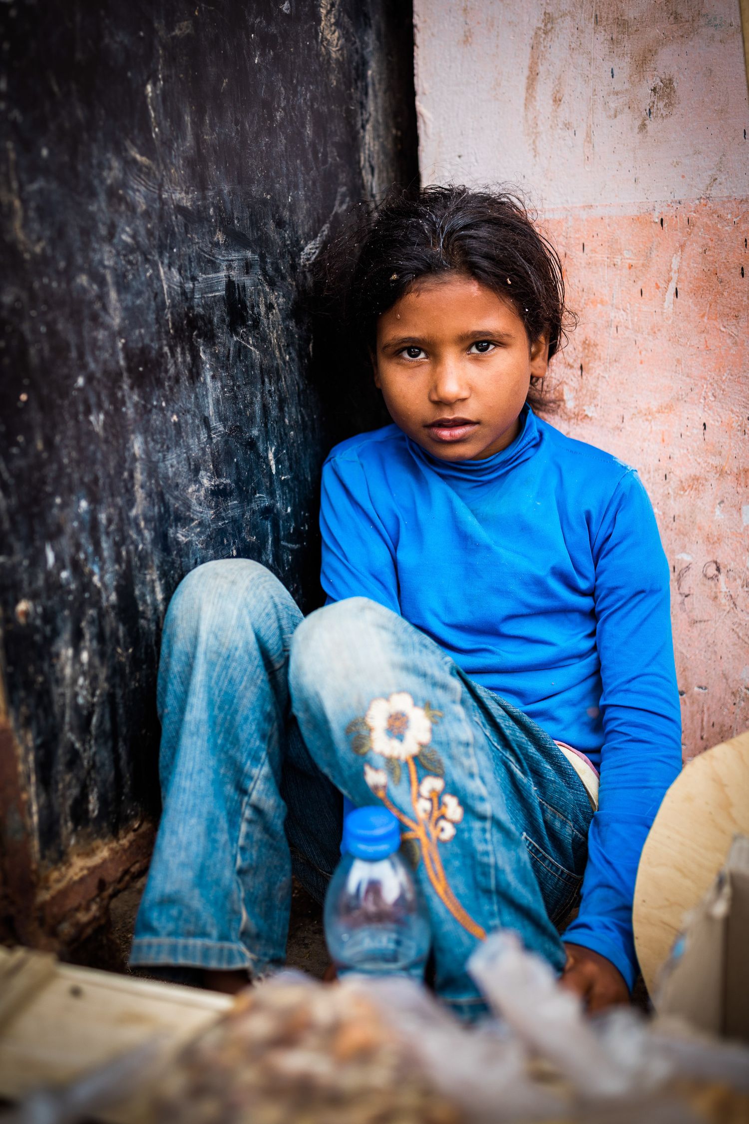A child in blue sits against a weathered wall with a water bottle nearby.