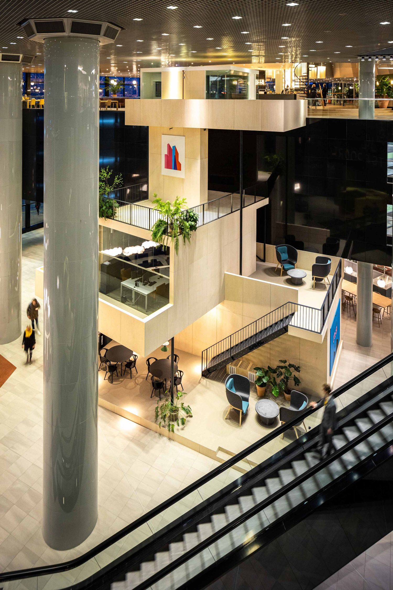 Modern multi-level shopping mall interior with escalators, columns and seating areas illuminated by warm overhead lighting.