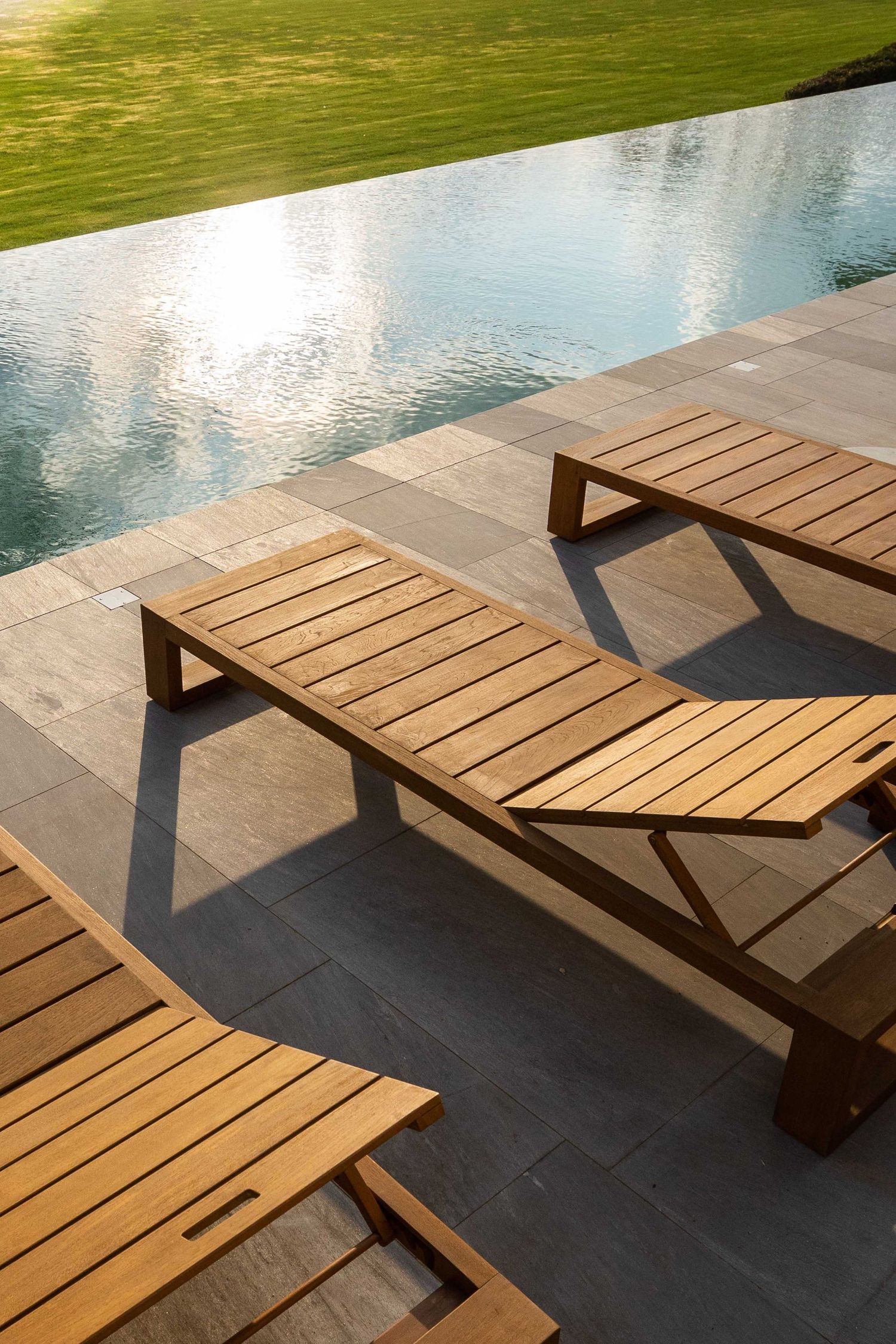 Wooden lounge chairs arranged along a poolside patio with shadows cast on dark stone tiles.