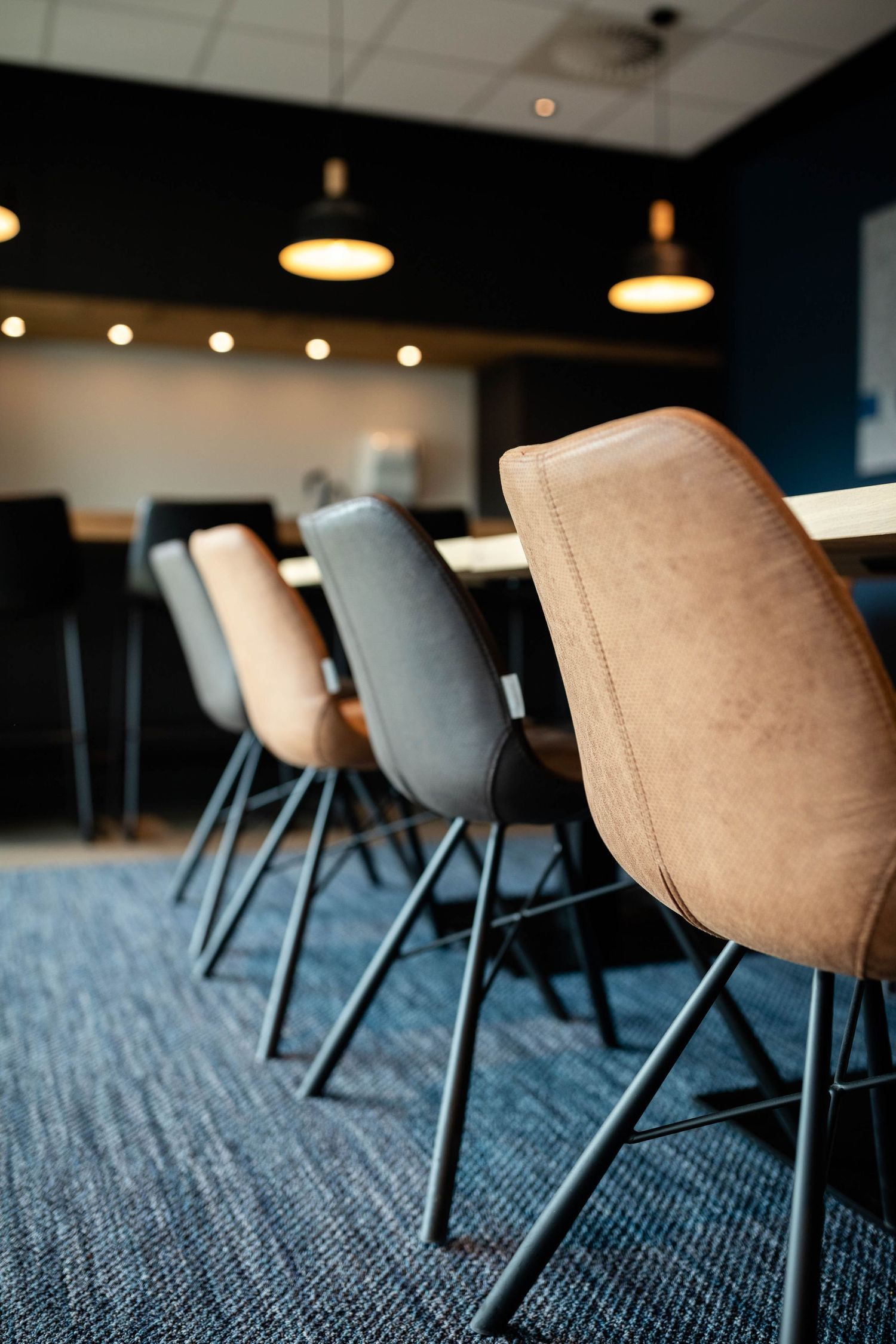 Modern office chairs in beige and gray tones arranged at a conference table on blue carpet with pendant lighting above.