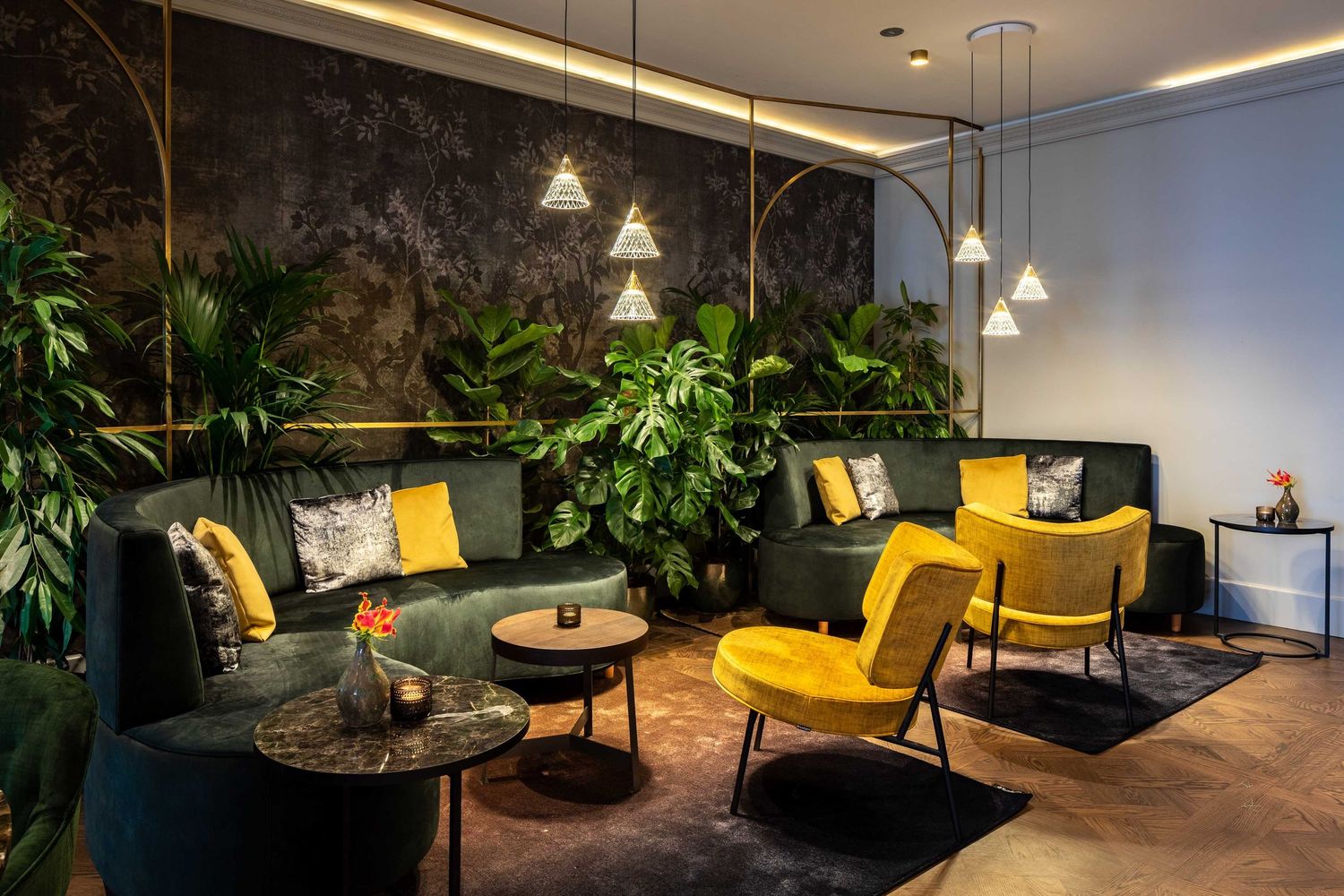 Modern office lounge area with yellow accent chairs, hanging pendant lights, and lush green plants creates a cozy workspace.