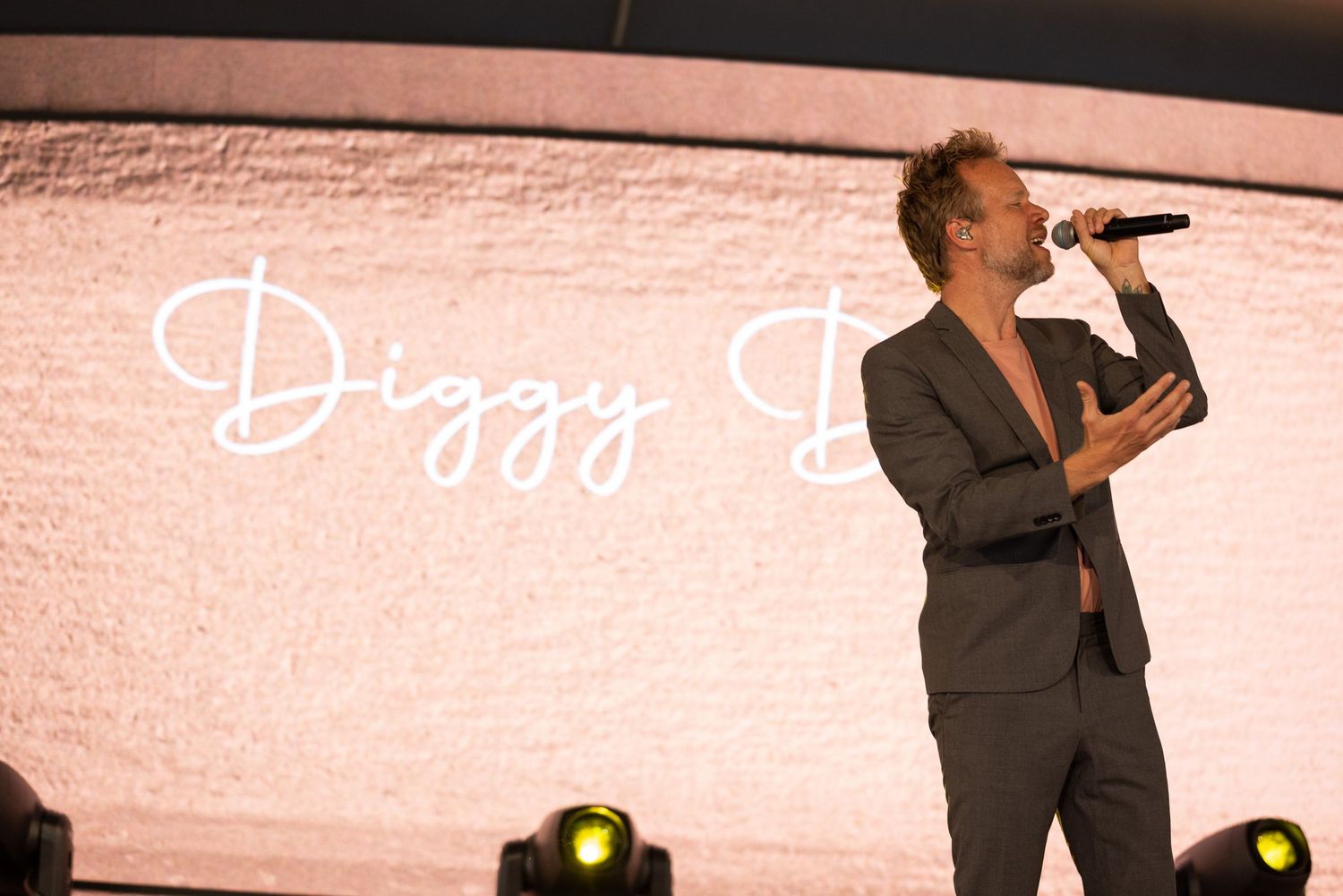 A performer in a gray suit sings passionately on stage with dramatic lighting effects in the background.