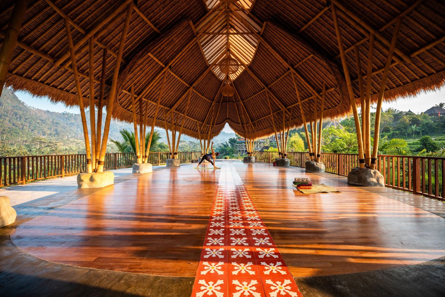 Interior view of a spacious bamboo yoga studio with traditional thatched roof and red patterned floor cushions.