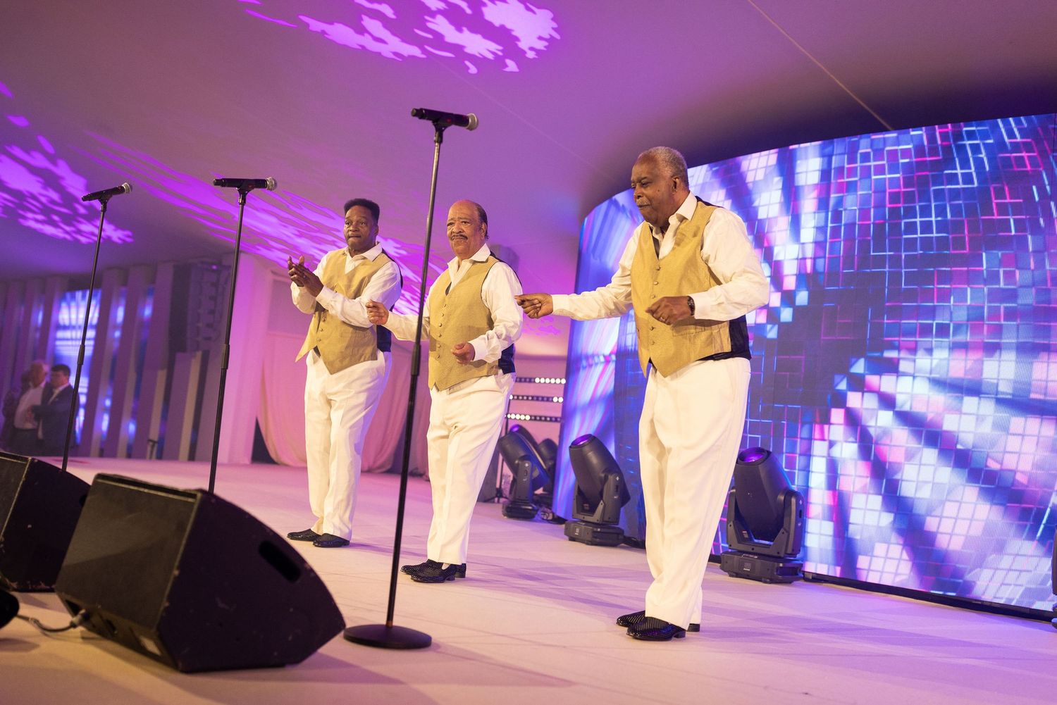 Performers in white suits singing on stage with purple and blue lighting effects in the background.