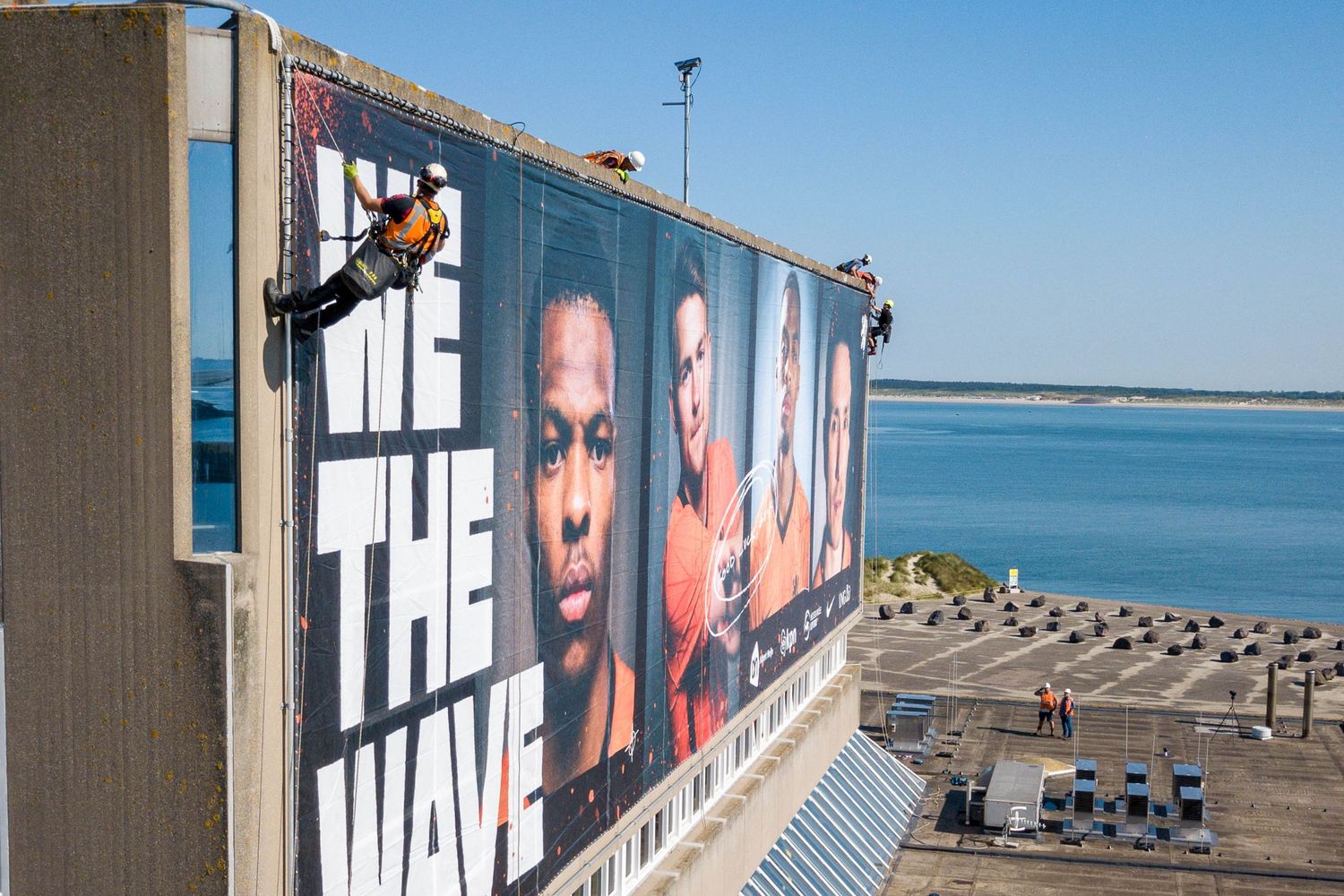 Workers rappelling down building installing large We The Wave Dutch football team banner.