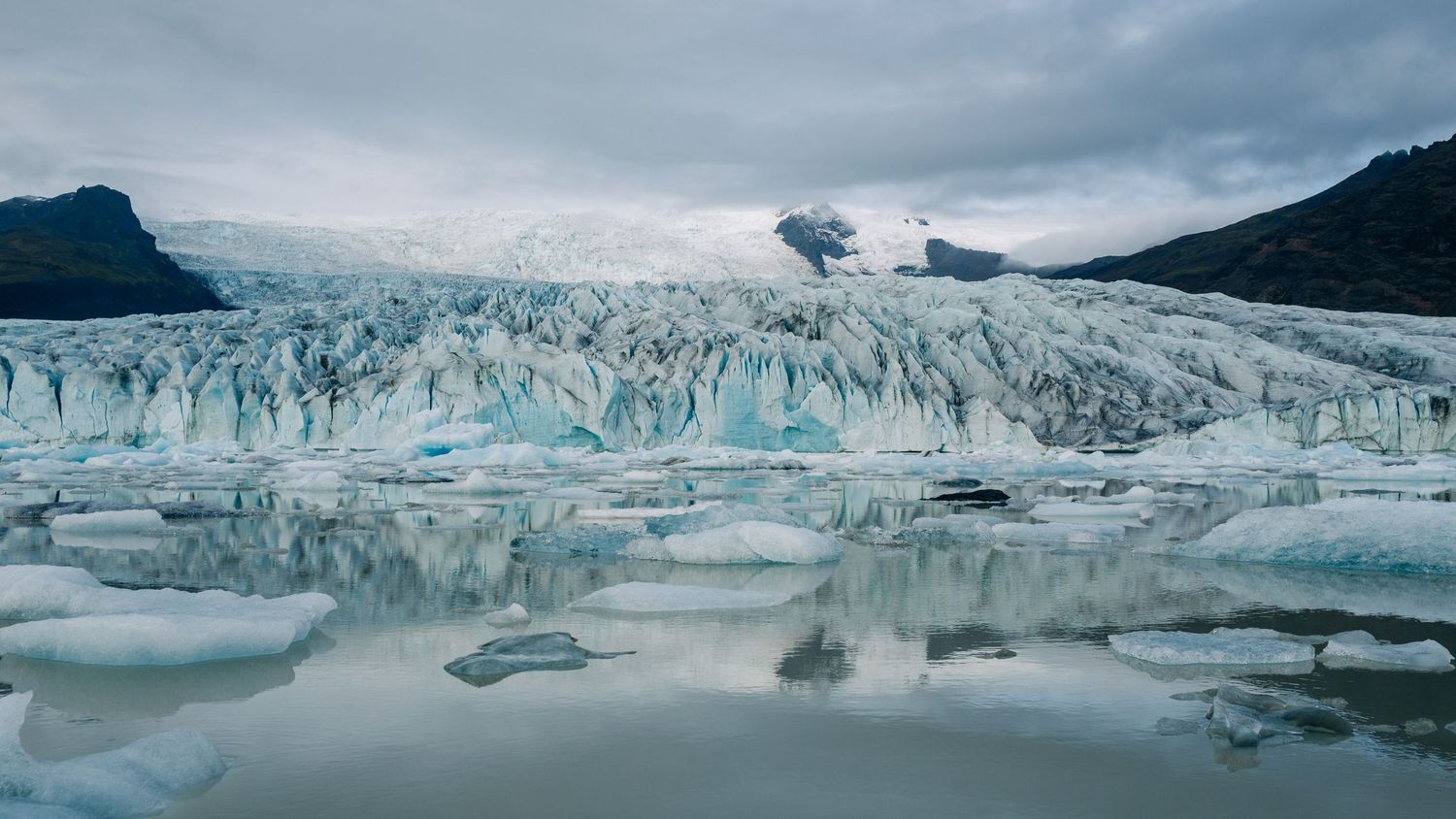 Panoramic view of a massive glacier with blue ice formations stretching across a dramatic cloudy landscape.