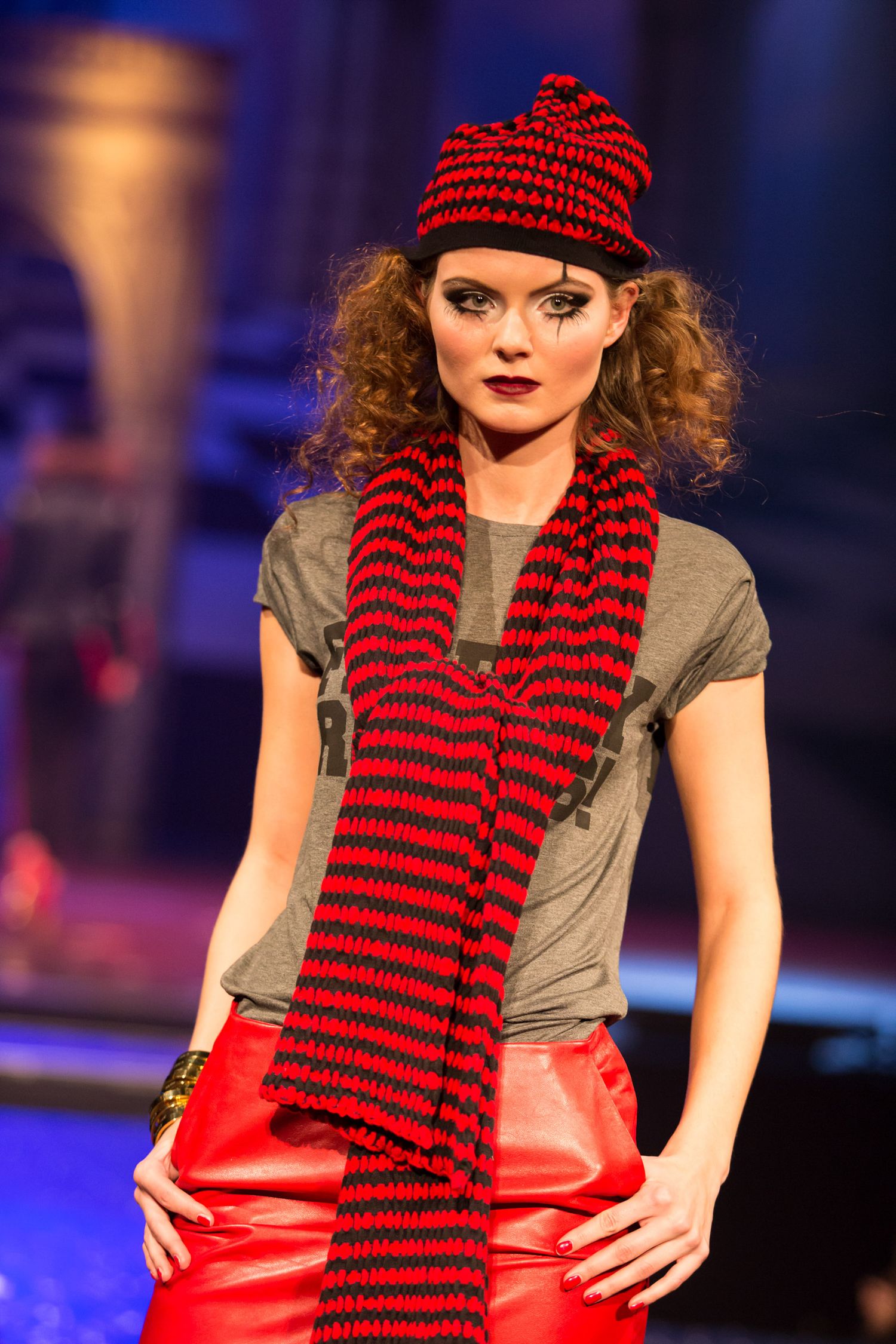 Model wearing red and black striped knit hat and scarf with gray tee and red leather pants on fashion runway.