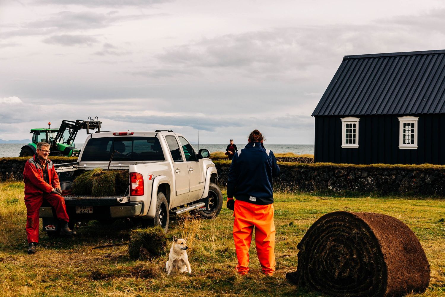 A series of small black houses with grass roofs dot the rugged Icelandic countryside under cloudy skies.