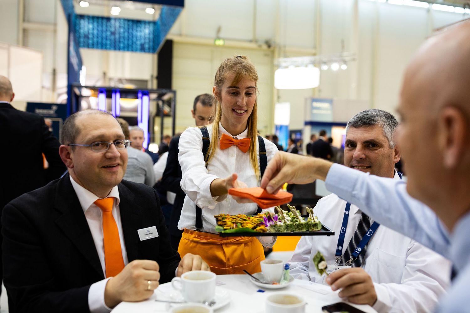A server in orange and white attire presents a tray of food to guests seated at a table during an event.