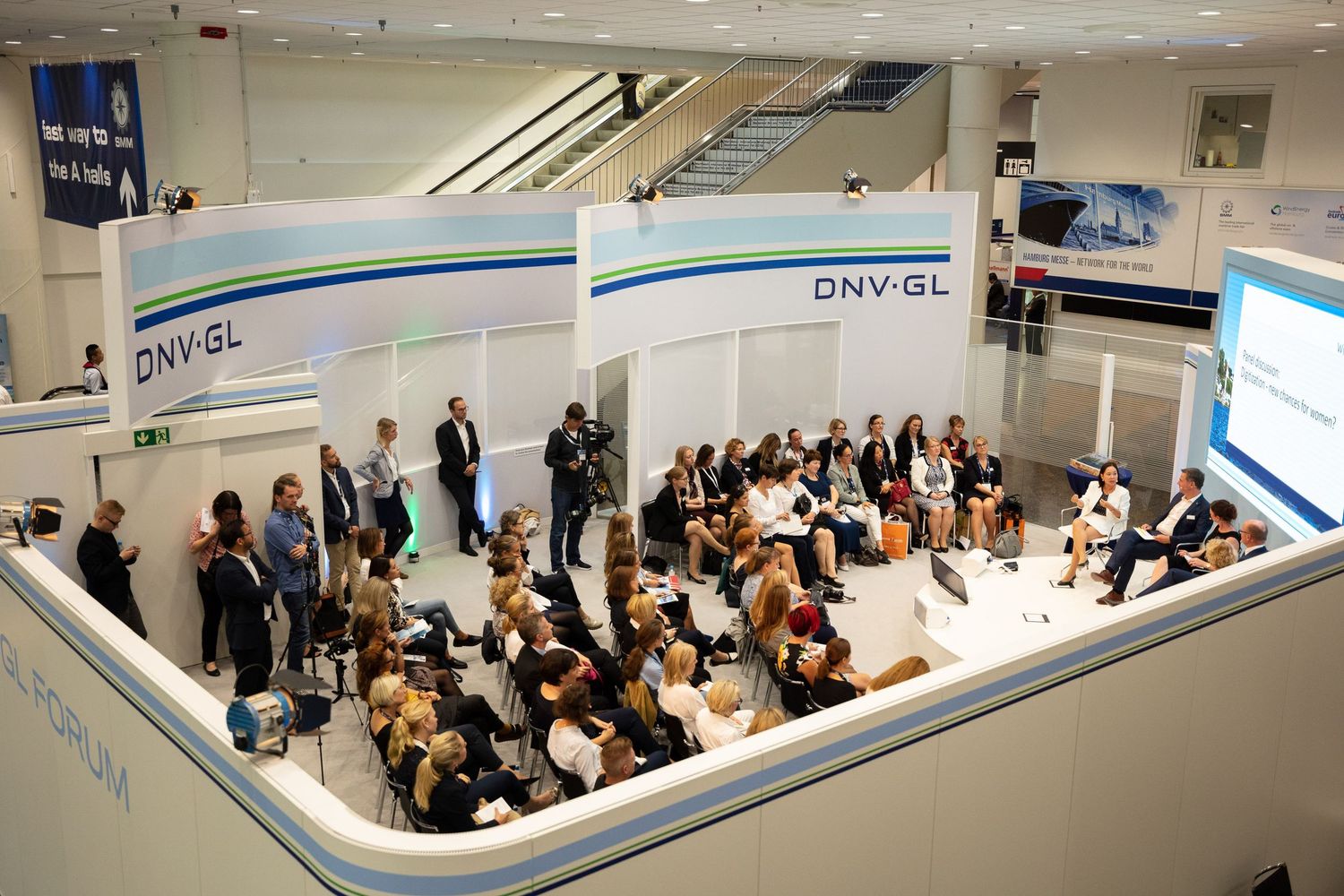 Business conference attendees seated in rows listen to a presentation in a modern white corporate meeting room.