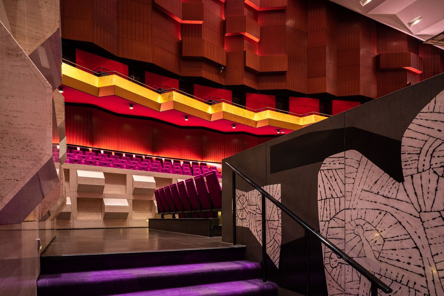 Modern theater corridor with angular walls features dramatic red and yellow lighting and geometric patterns on the wall.