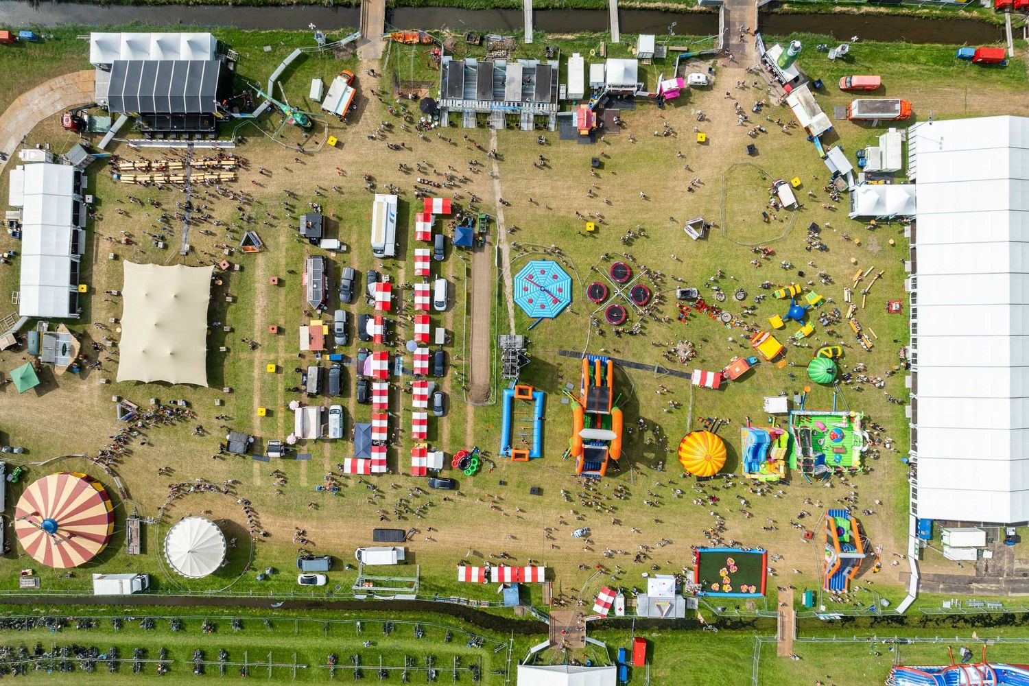 Aerial view of a colorful outdoor fairground with carnival rides, inflatable attractions, and crowds on green grass.