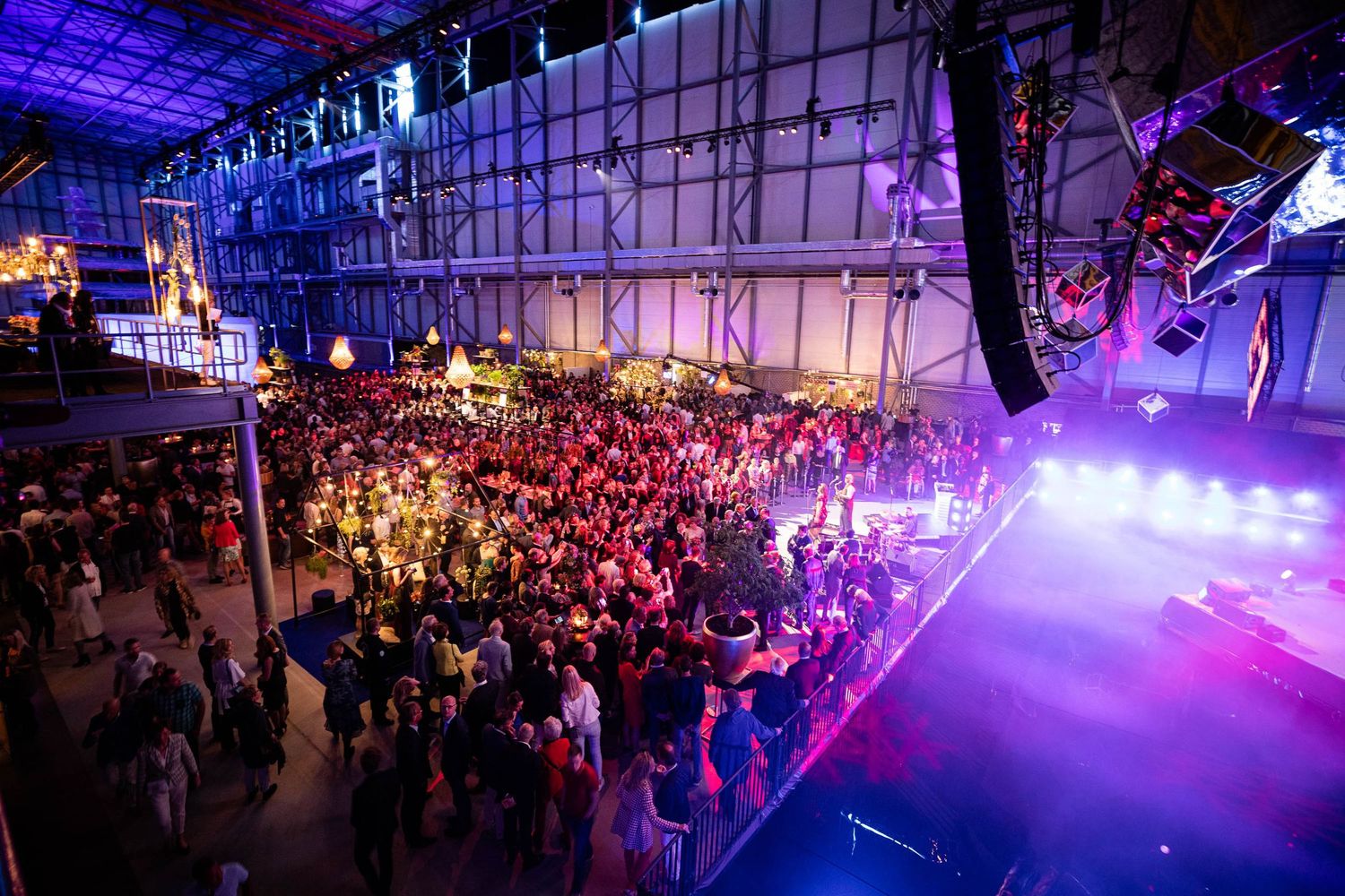 Panoramic view of a concert venue with purple lighting and fog effects illuminating a packed crowd of attendees.