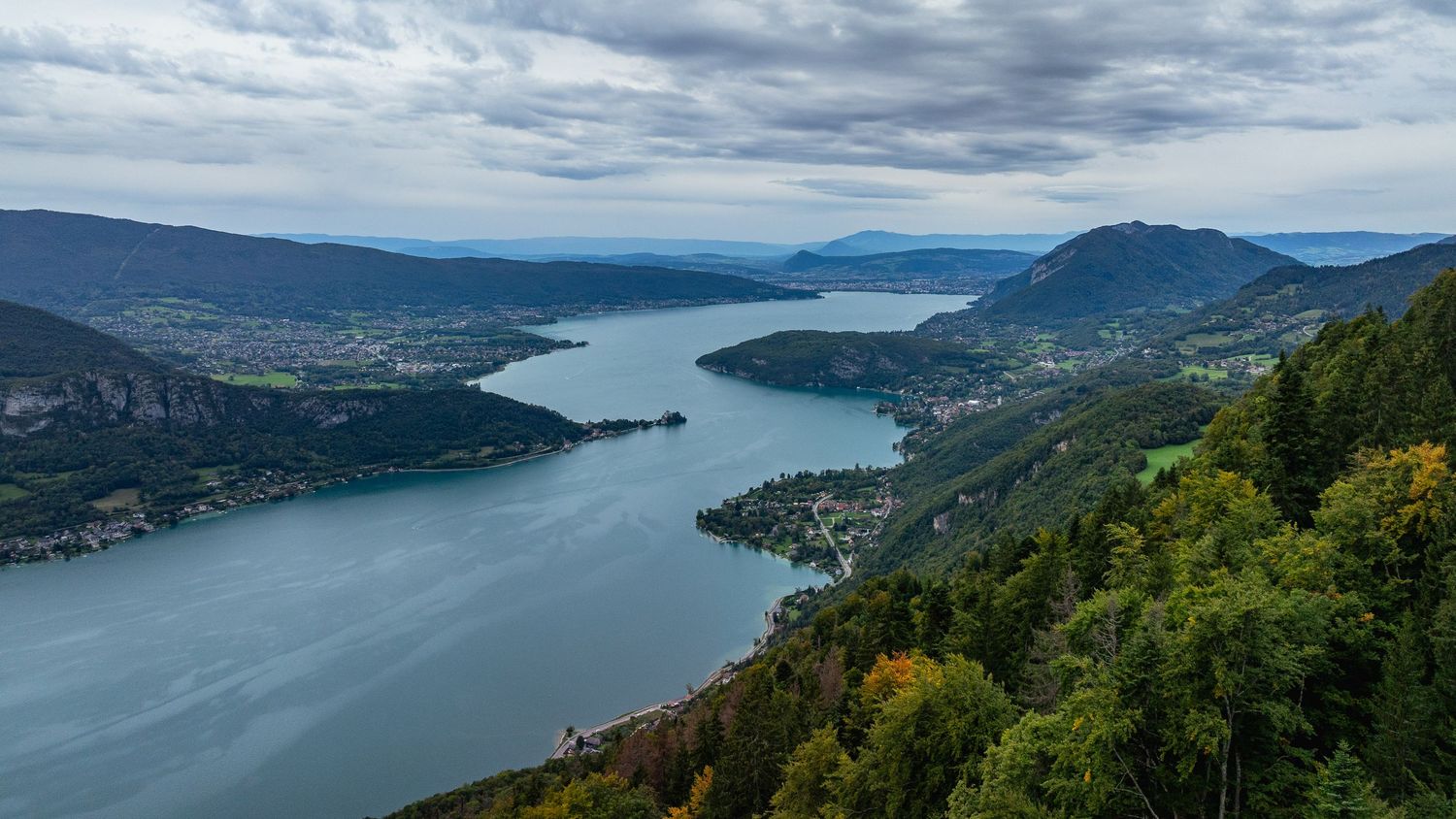 Wide panoramic view of a mountain lake valley with forested slopes and cloudy skies.