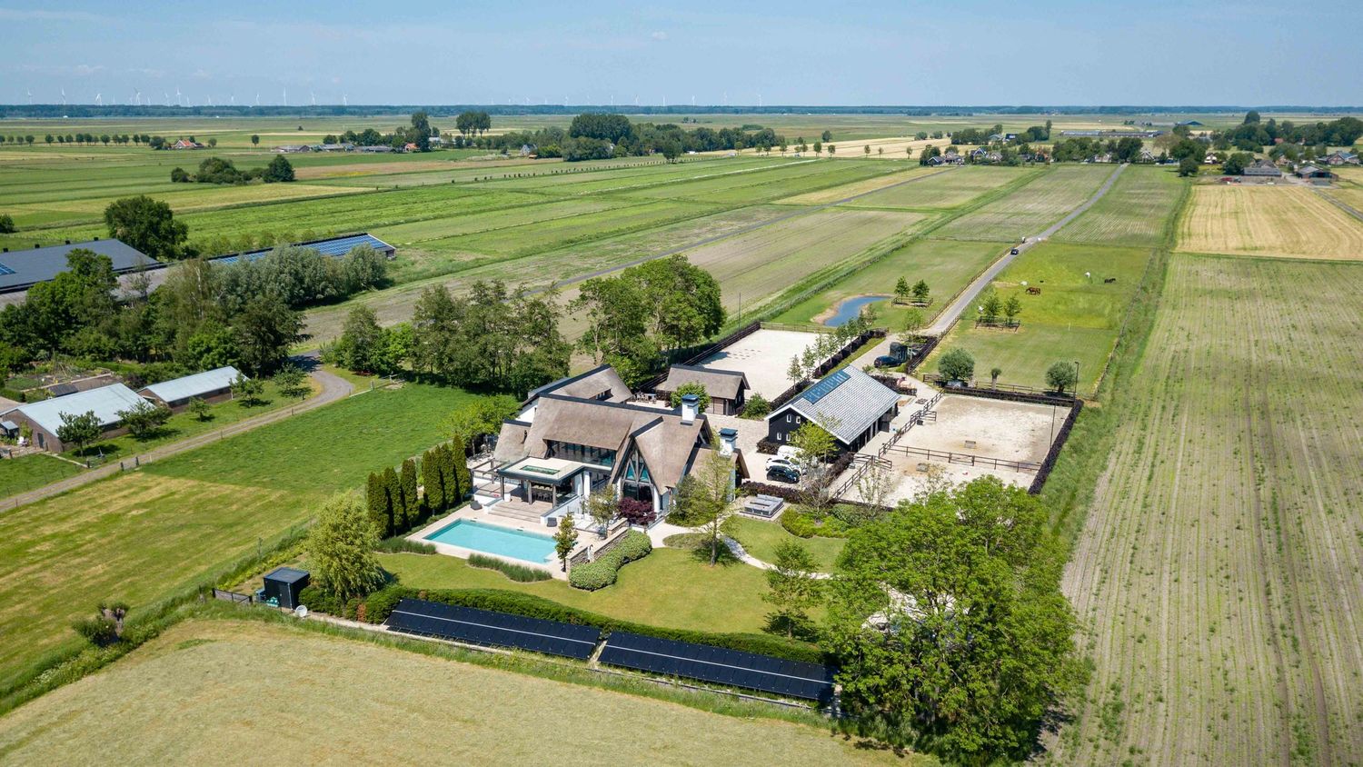Panoramic aerial view of a rural landscape with green fields, farm buildings, and swimming pools on a sunny day.