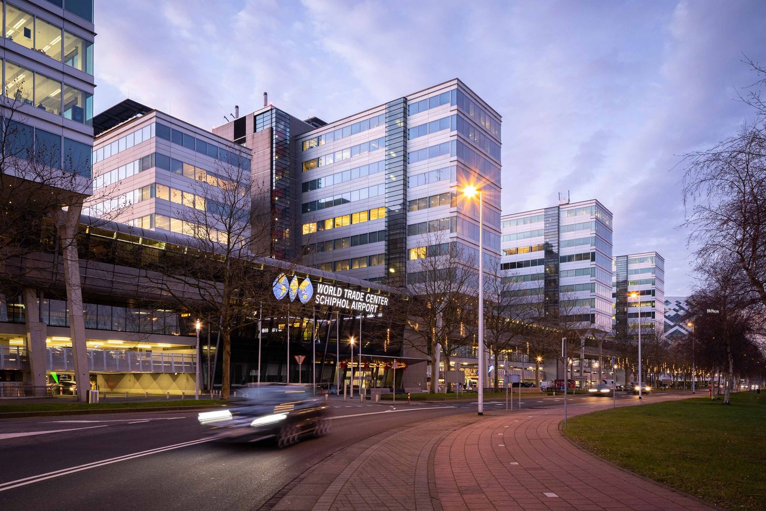 Modern office buildings with illuminated windows at dusk along a curved street with passing cars.