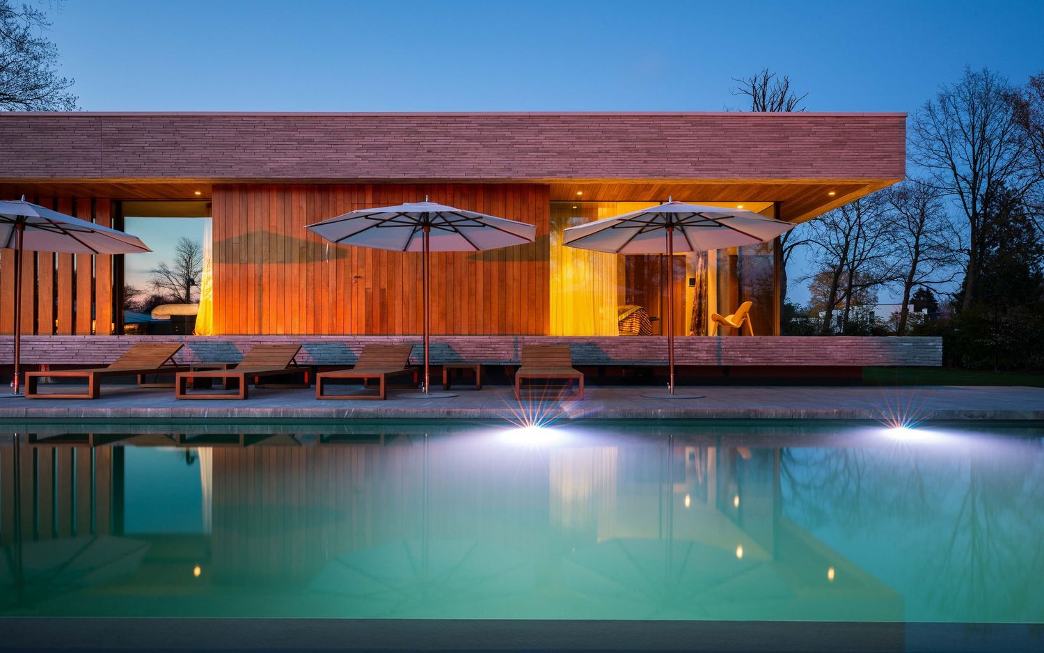 Illuminated outdoor swimming pool at dusk with glowing turquoise water and patio umbrellas by a modern building.