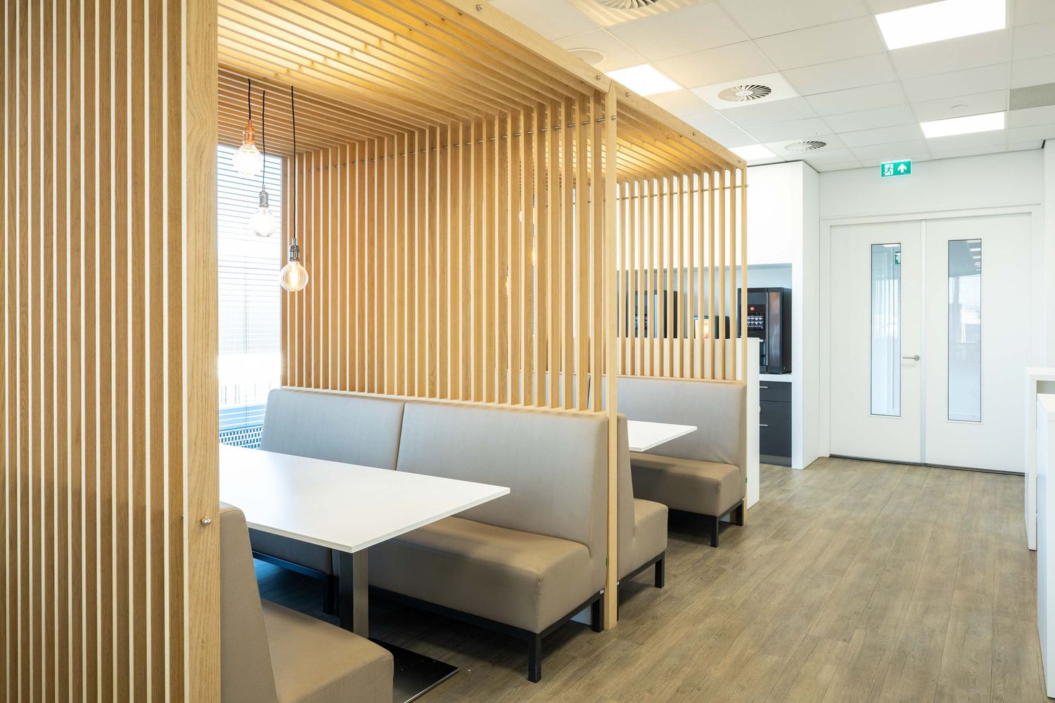 Modern office break room with wooden slat walls, gray booth seating, and hardwood floors in a minimalist design.