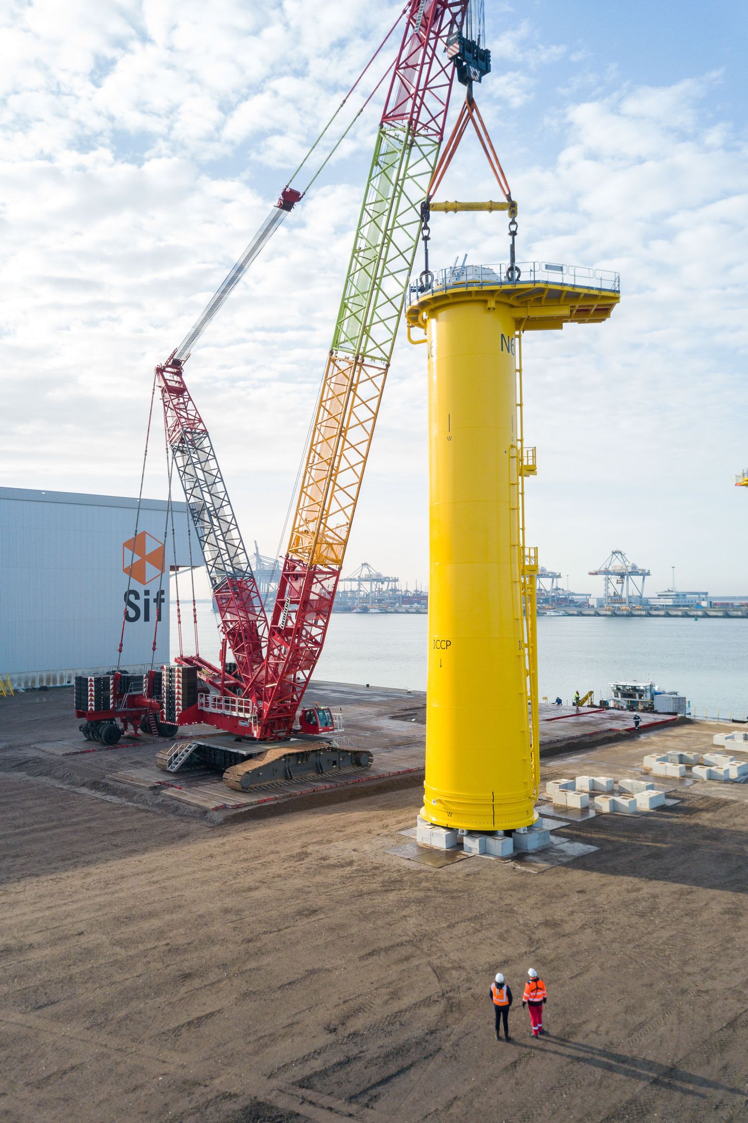 Yellow offshore wind turbine monopile being lifted by red crawler crane at industrial port with two workers observing.