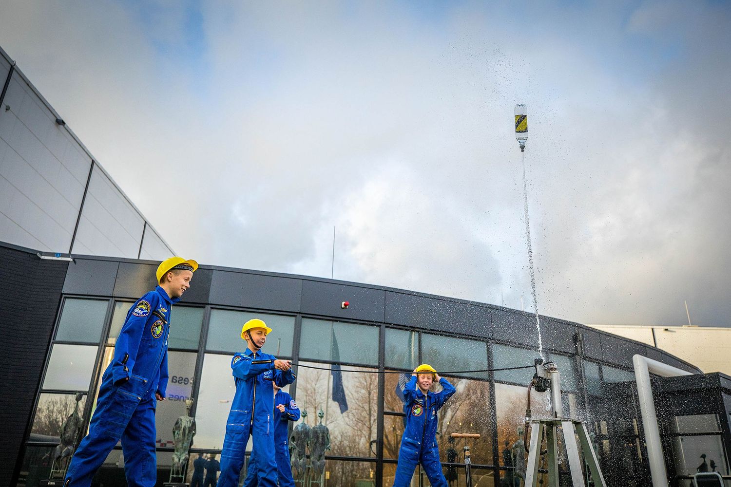 Three workers in blue overalls and yellow hard hats operating equipment outside a modern building.