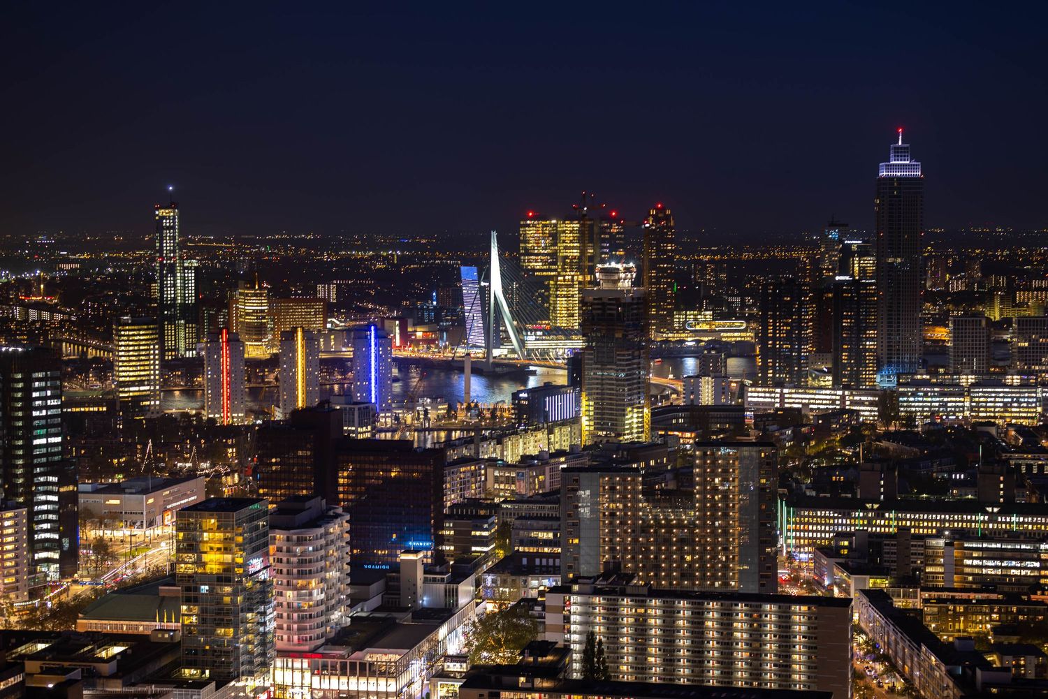 Aerial night view of Rotterdam cityscape with illuminated skyscrapers and the iconic Erasmus Bridge glowing colorfully.
