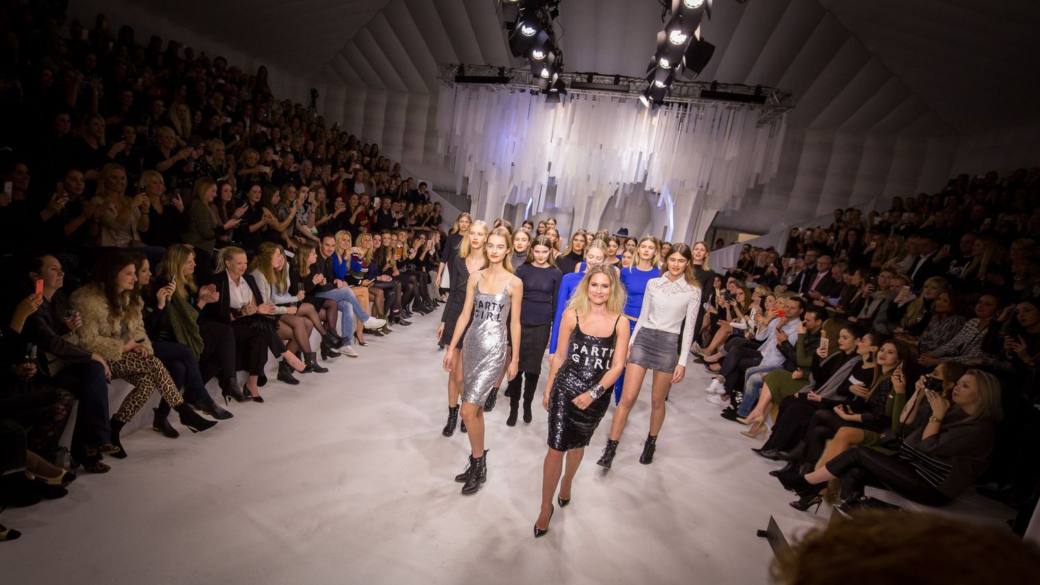 Models walk the runway at a glamorous fashion show with a packed audience under dramatic lighting.