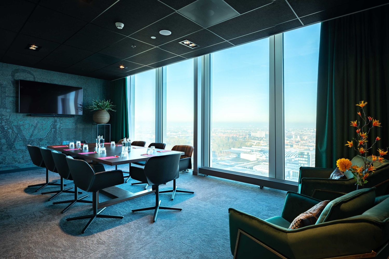 Modern conference room with floor-to-ceiling windows offering city views, featuring sleek furniture and floral decor.