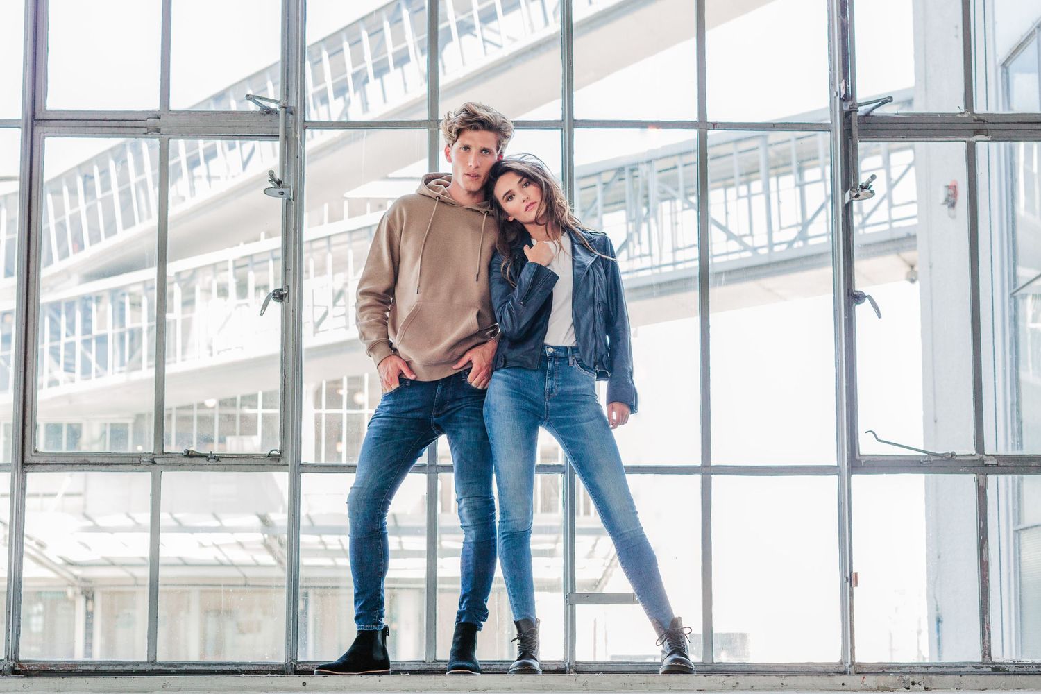 Fashion lifestyle photoshoot of couple posing casually by large industrial windows.