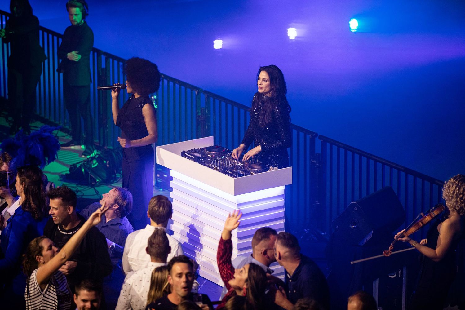 A DJ performs at an elevated booth with blue and purple stage lighting while crowd members dance below.