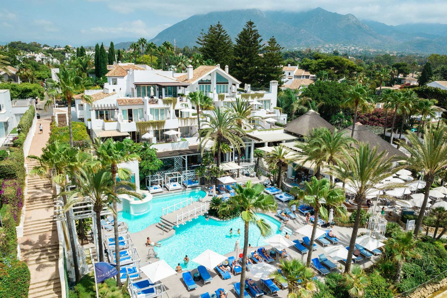 Aerial view of a luxury resort with a sparkling pool, blue loungers, palm trees, and mountain backdrop in Marbella.