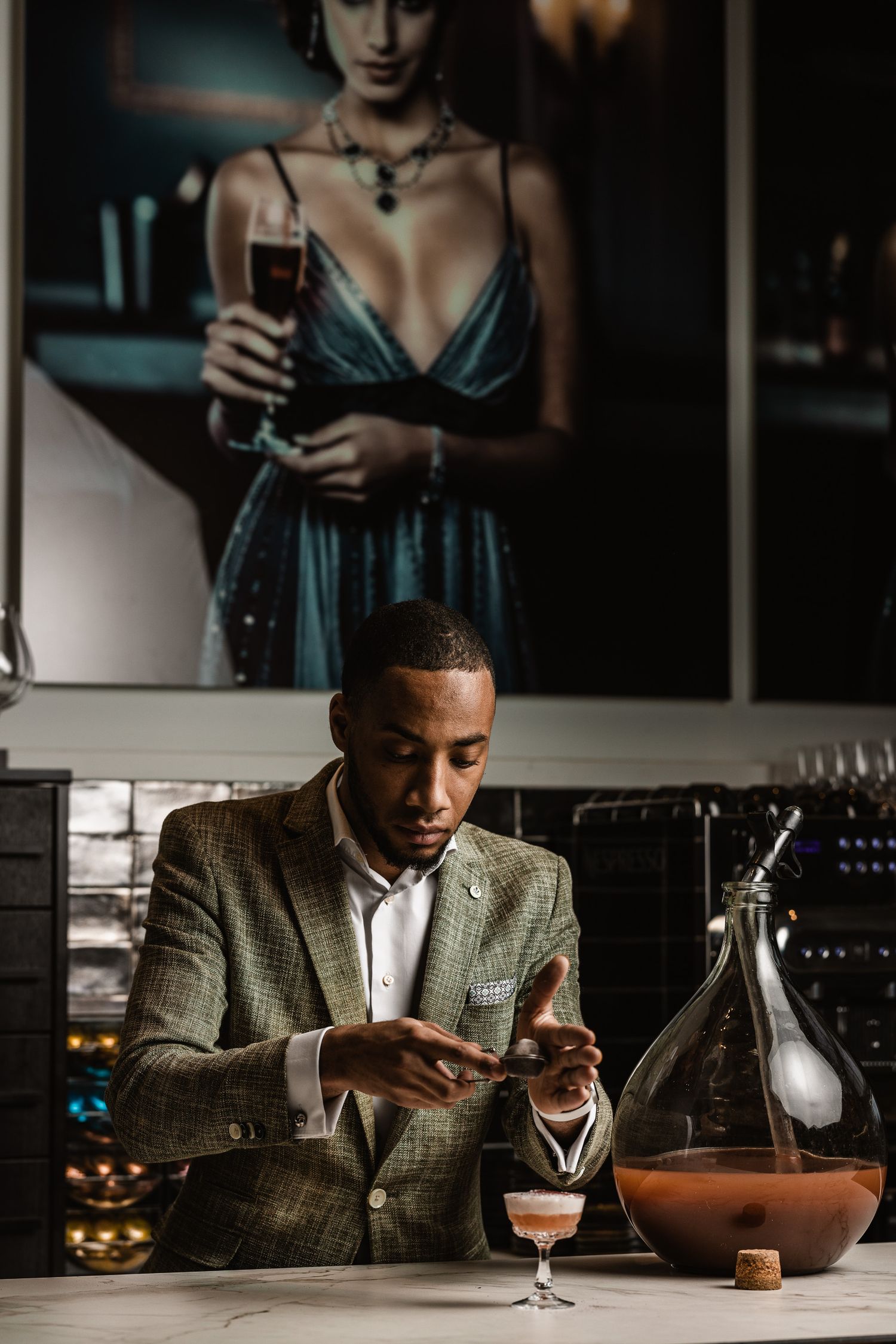 A person in a tweed blazer sits at a dimly lit bar examining their phone and drink in an atmospheric setting.