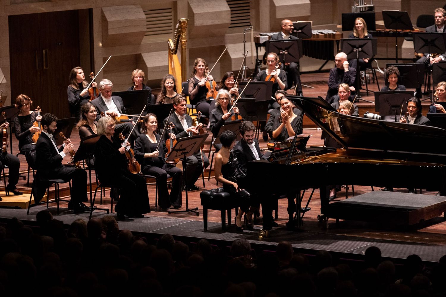 Symphony orchestra performs on stage with grand piano and string section during classical concert performance.