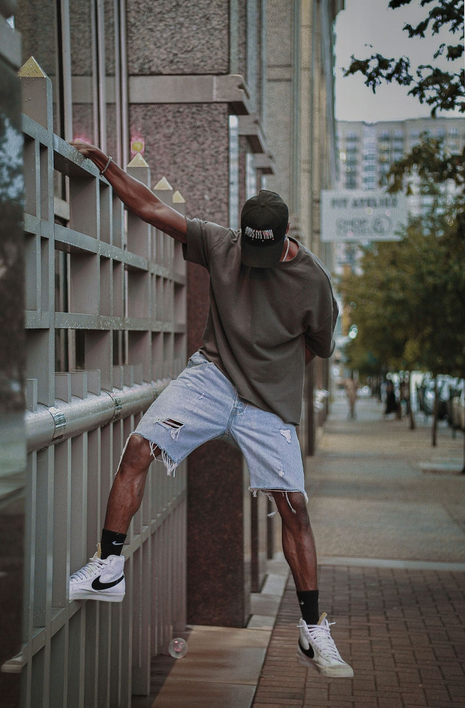 A young man in ripped denim shorts, olive tee, and Nike sneakers balances on urban railings in a city setting.