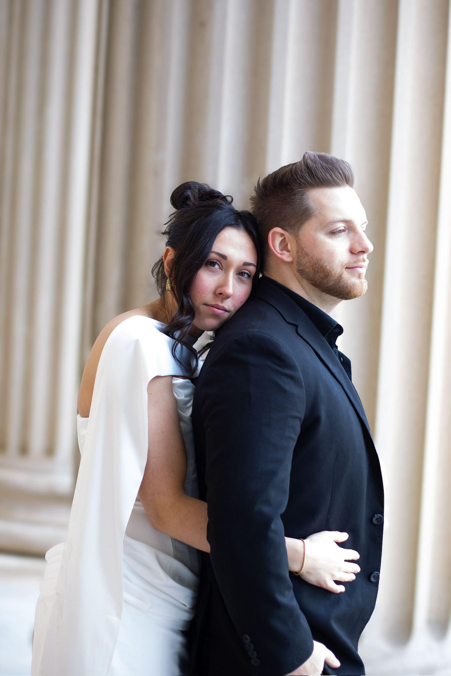 Bride in white dress embraces groom in black suit, leaning against classical columns in elegant wedding portrait.