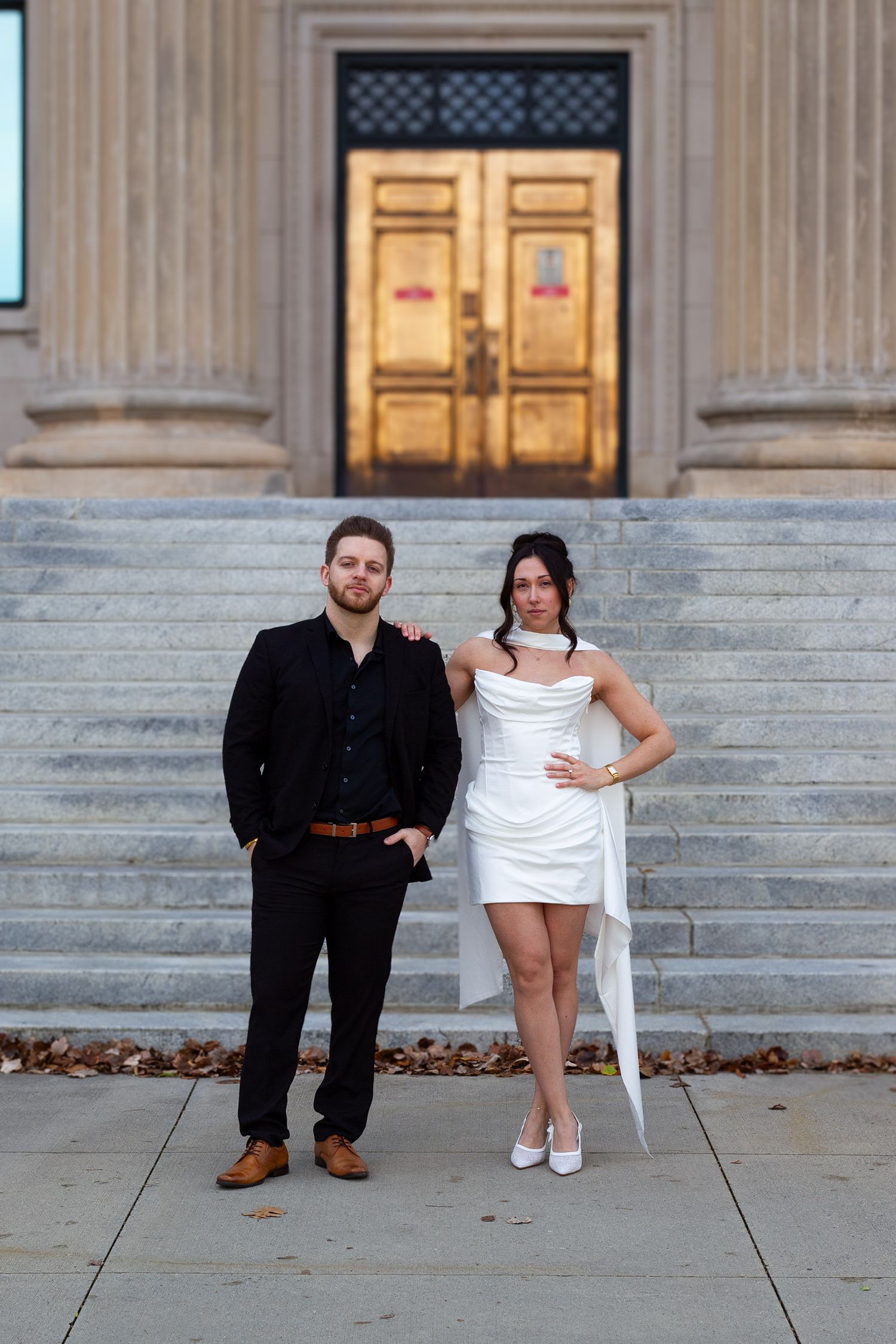 Couple in wedding attire posing on steps of a grand building, groom in black suit, bride in short white dress.
