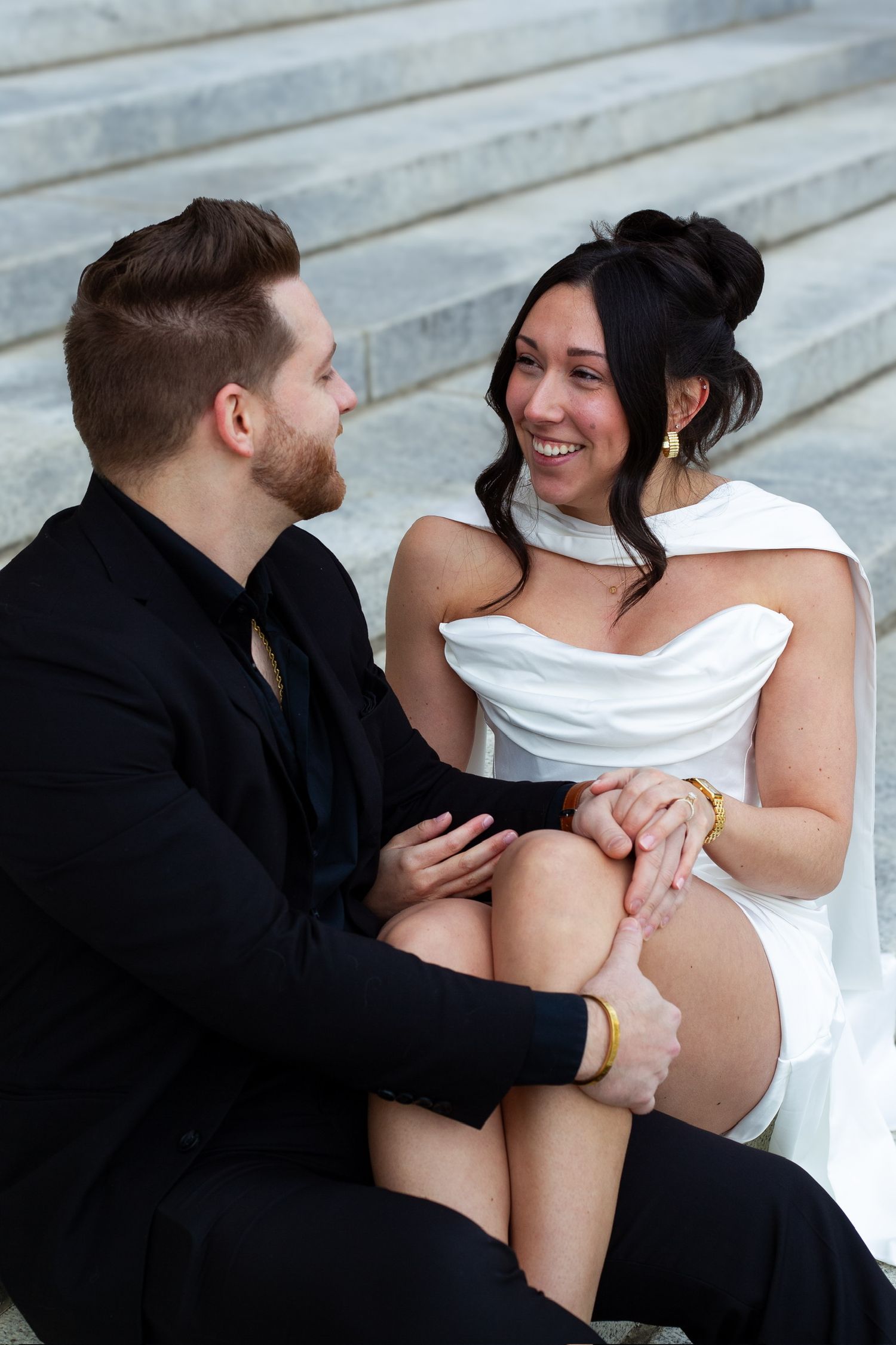Happy bride in white dress and groom in black suit sitting on stone steps, holding hands and smiling at each other.