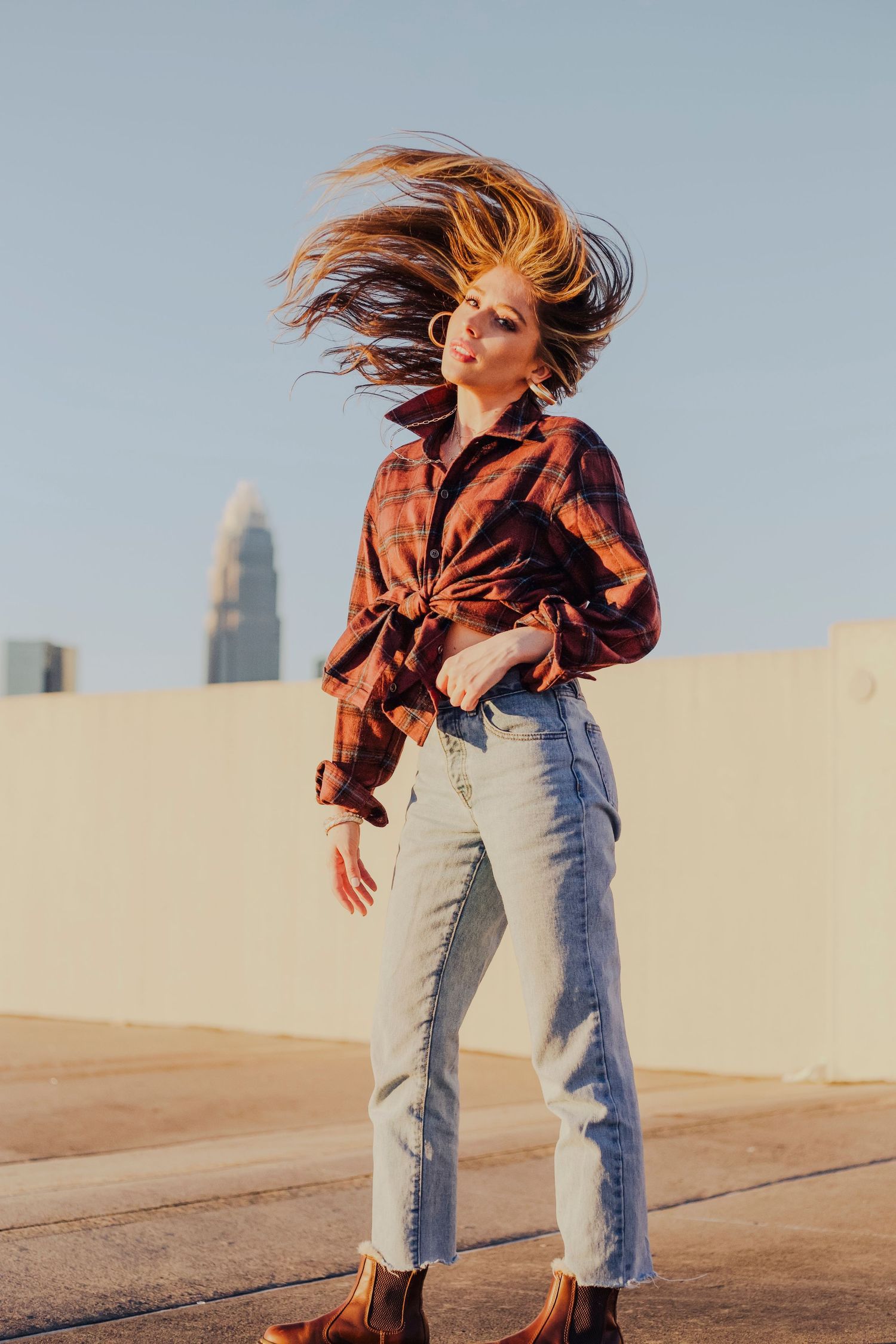 Woman in plaid shirt and jeans with hair flipping on rooftop against city skyline backdrop.