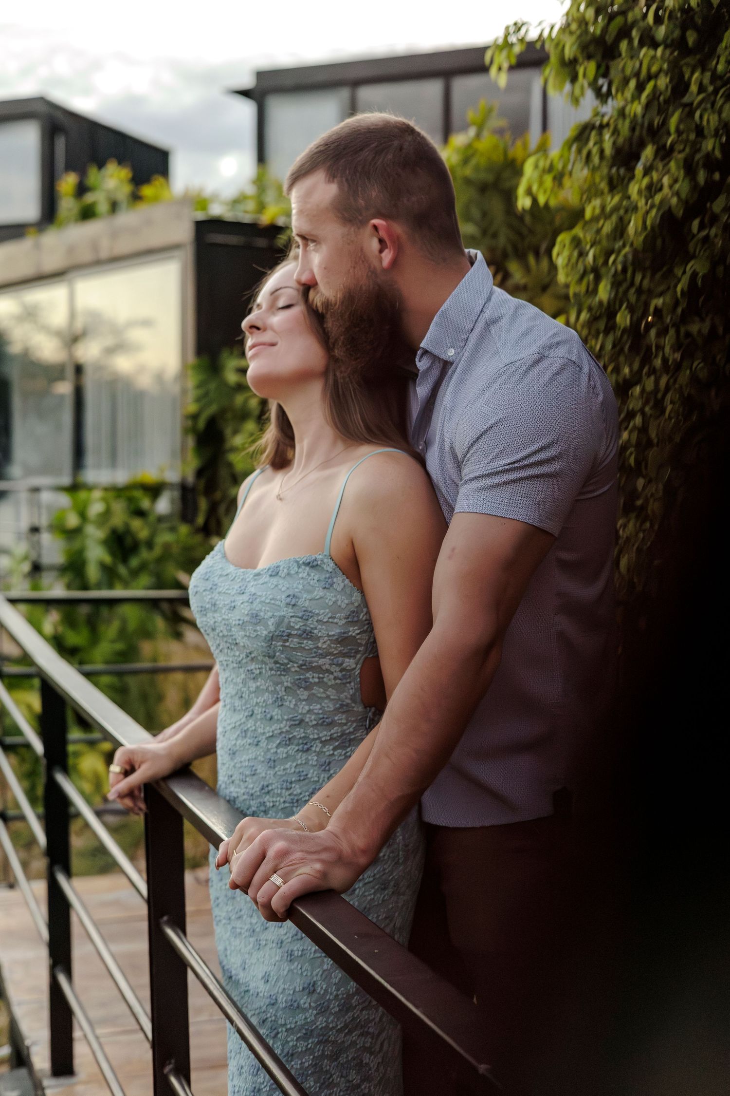 Romantic couple embracing on a balcony, man kissing woman's head, surrounded by lush green foliage.