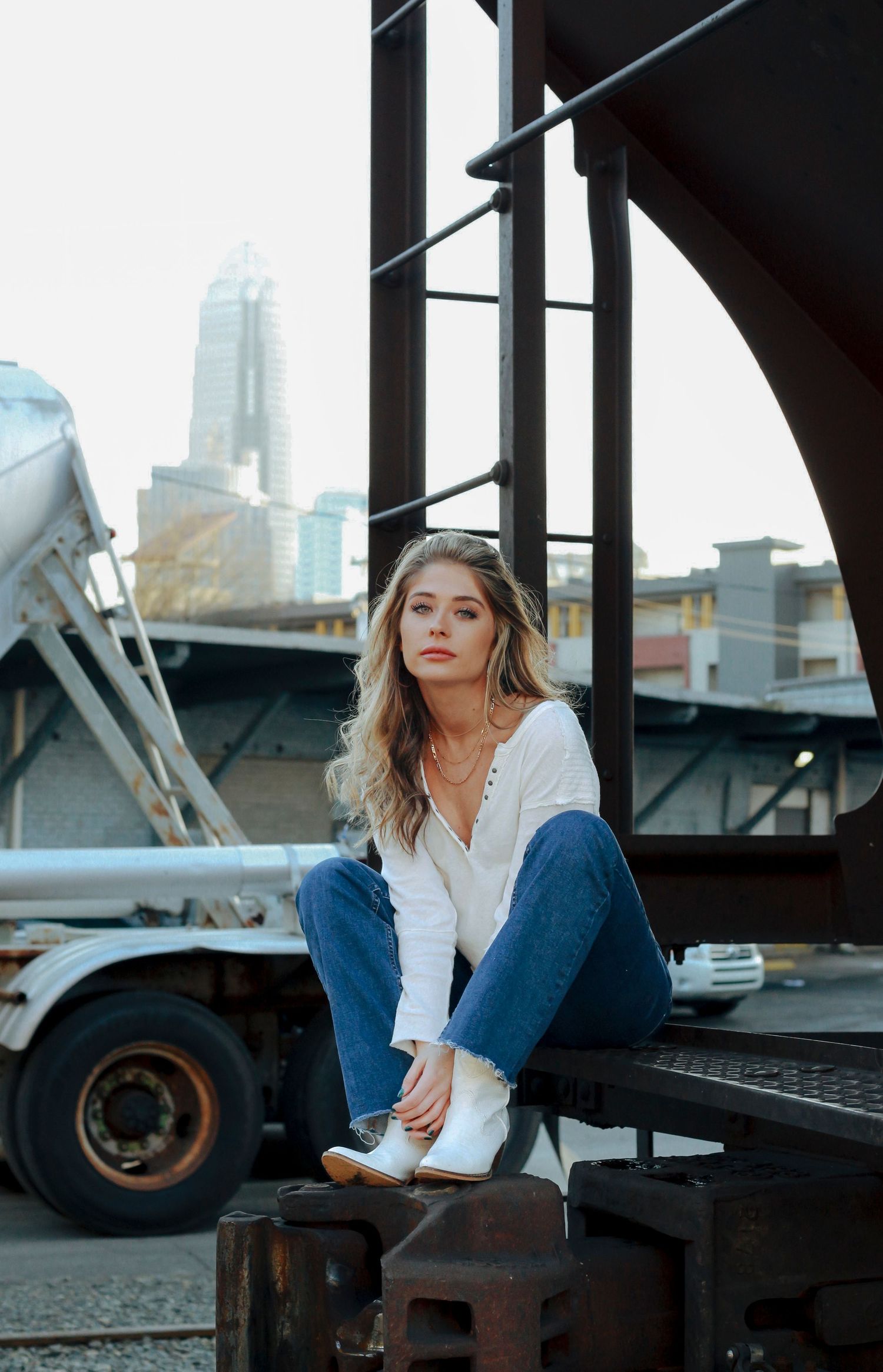 Young woman in white top and jeans sitting on rustic metal structure with city skyline in background.