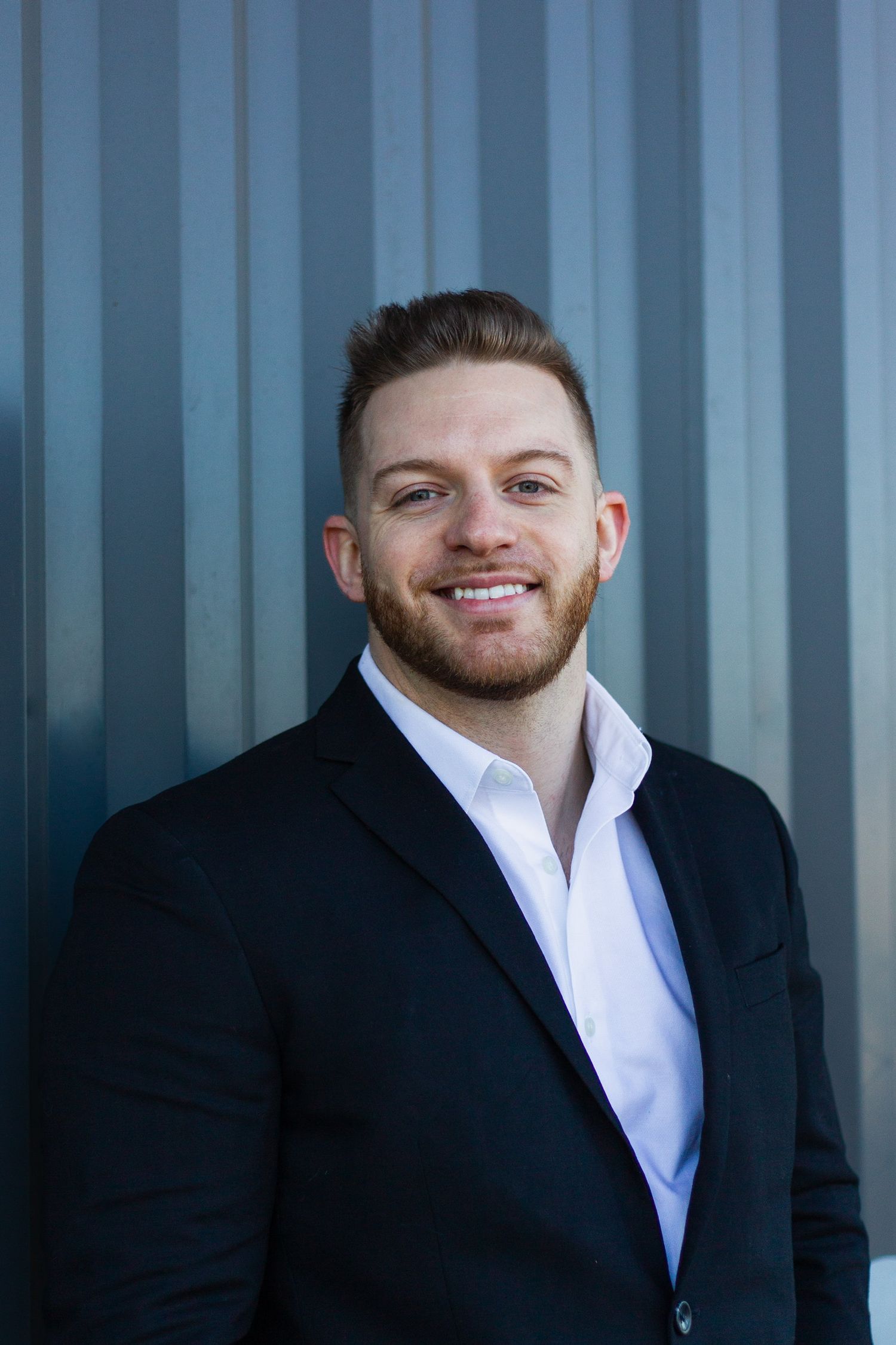 Smiling young businessman with beard wearing black suit and white shirt poses against gray metal wall.