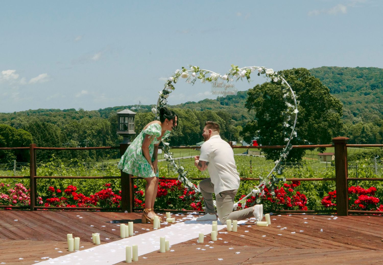 Man proposing to woman on decorated outdoor deck with heart-shaped floral arch, candles, and lush green vineyard backdrop.